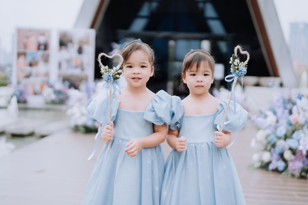 Two little flower girls in matching light blue dresses hold decorative wands at an outdoor wedding ceremony.