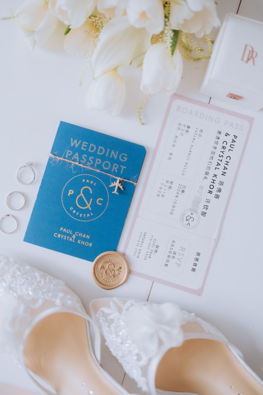 A wedding passport, plane ticket, and elegant white bridal shoes arranged with delicate white flowers and rings.
