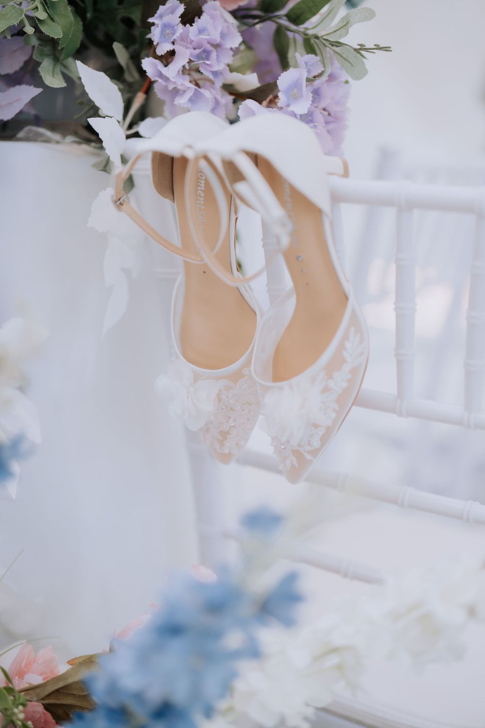 Delicate white lace wedding shoes with soft purple flowers in the background create an elegant bridal detail.
