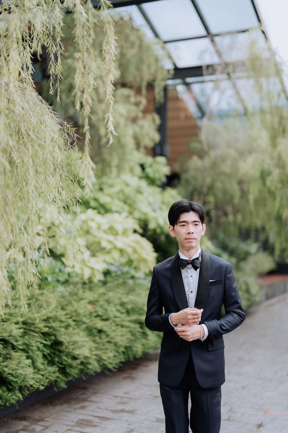 A person wearing a black tuxedo with bow tie poses elegantly in front of hanging willow branches in a garden setting.