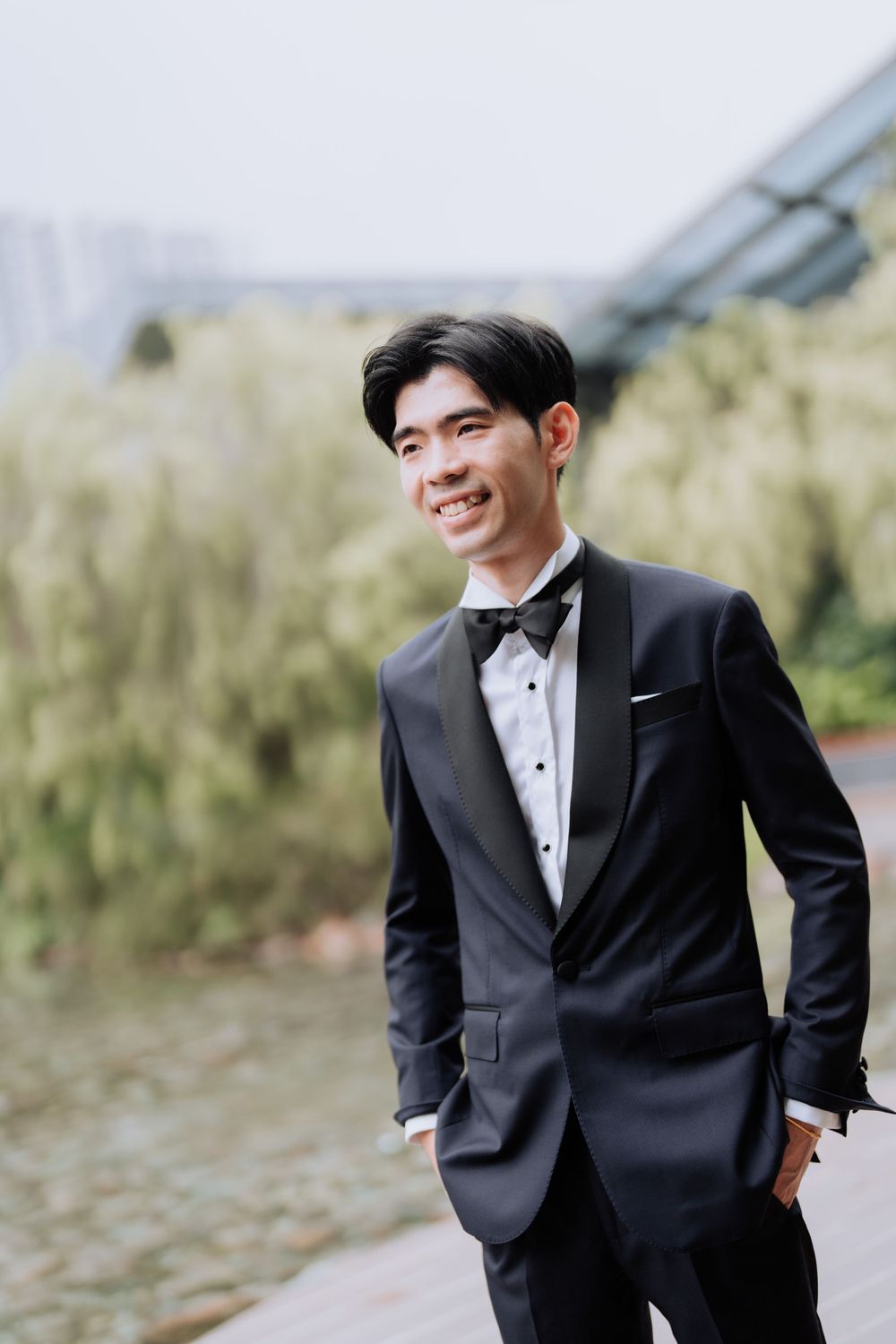 A groom in a classic black tuxedo and bow tie smiles while standing outdoors.