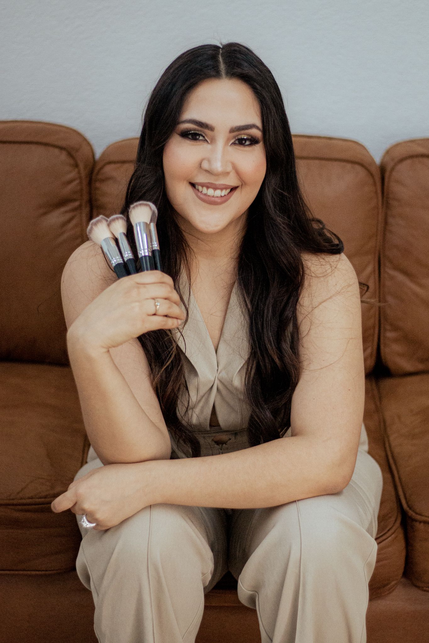Professional holding makeup brushes sits smiling on brown leather couch in beige outfit.