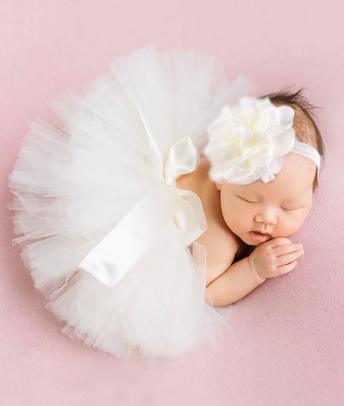 Sleeping newborn in white tutu and headband on pink background.