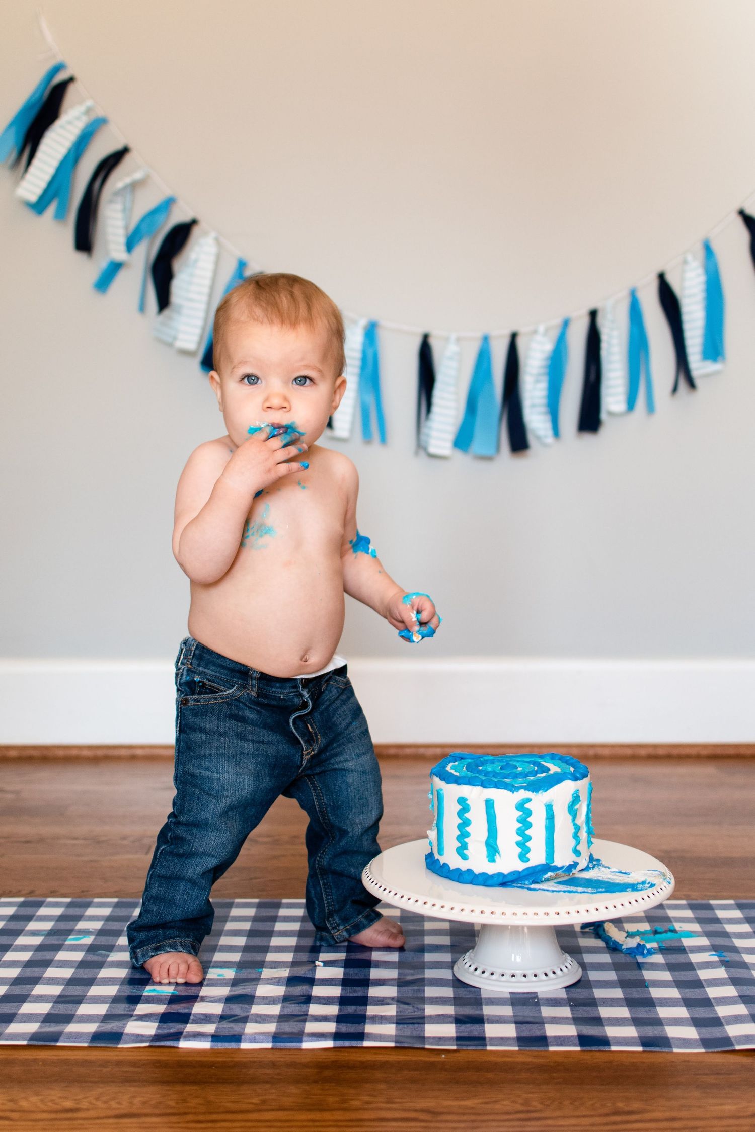 A baby stands on a checkered blanket next to a blue and white smash cake with matching garland decorations in the background.