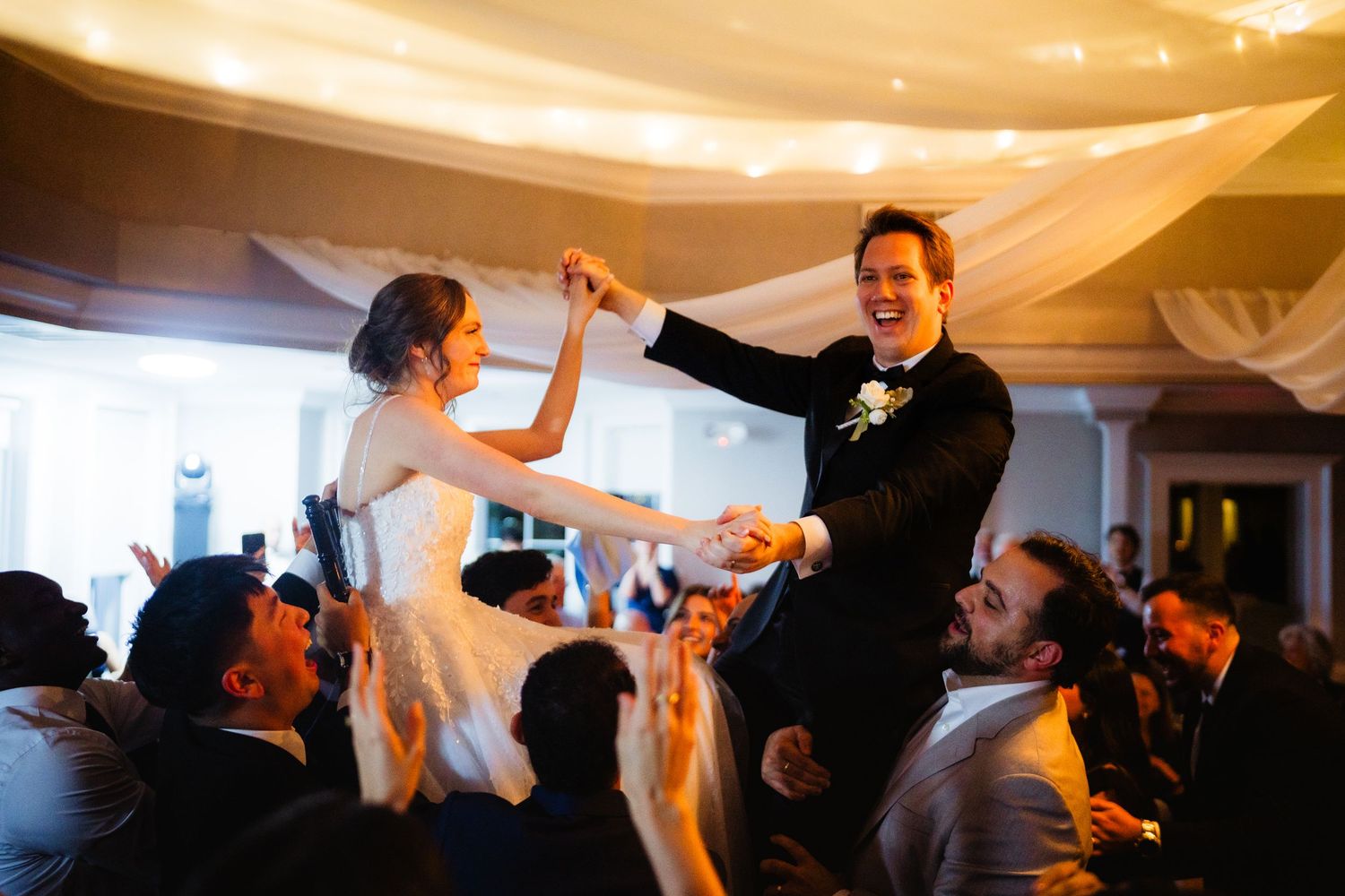Bride and groom dancing joyfully on guests shoulders at elegant wedding reception with draped ceiling.
