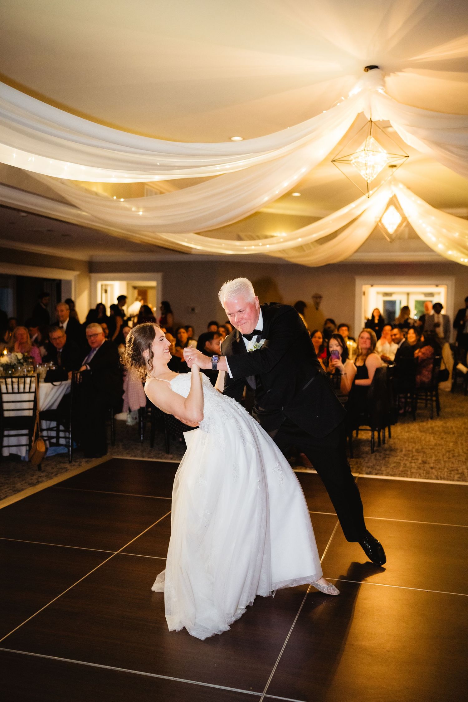 Bride and father share elegant first dance at wedding reception with draped ceiling decor and guests watching.
