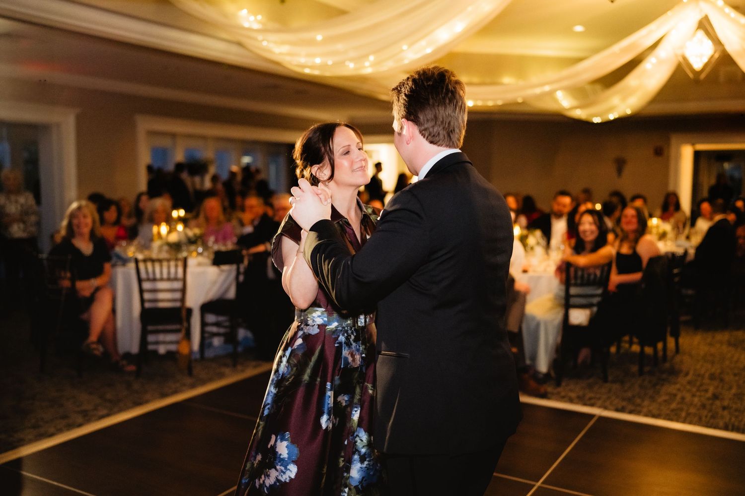 Couple shares first dance at elegant wedding reception with guests watching under draped ceiling lights.
