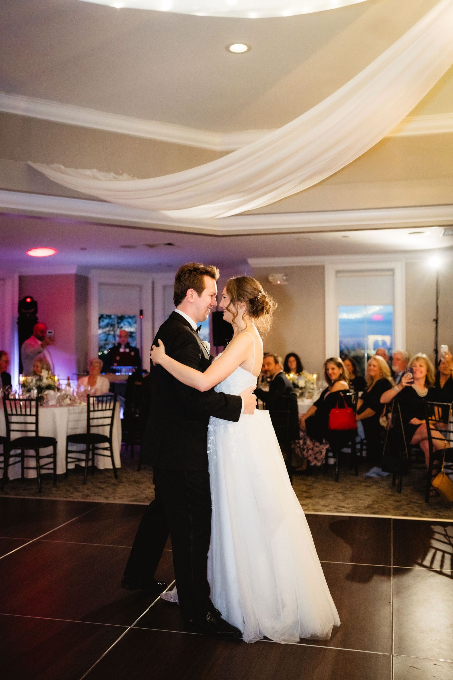 Bride and groom share their first dance at elegant wedding reception with draped ceiling and guests watching.