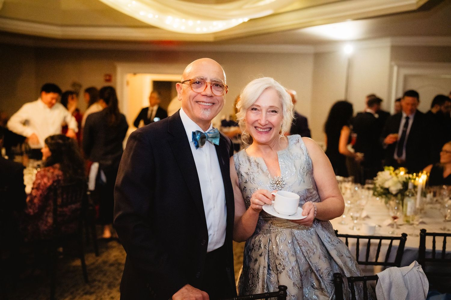 Elderly couple smiling at a formal wedding reception, man in bow tie, woman holding a teacup.