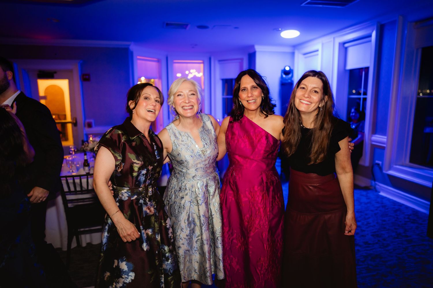 Four women in elegant formal dresses pose together smiling at an evening wedding reception with blue ambient lighting.