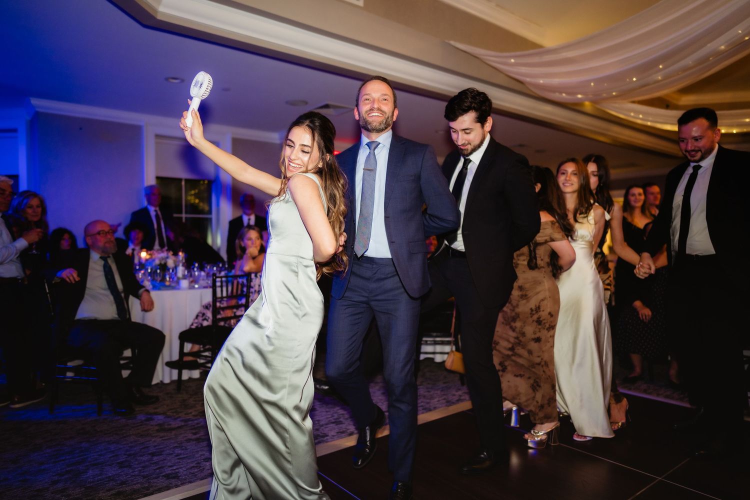 Guests dancing joyfully at an elegant wedding reception with blue ambient lighting and formal attire.