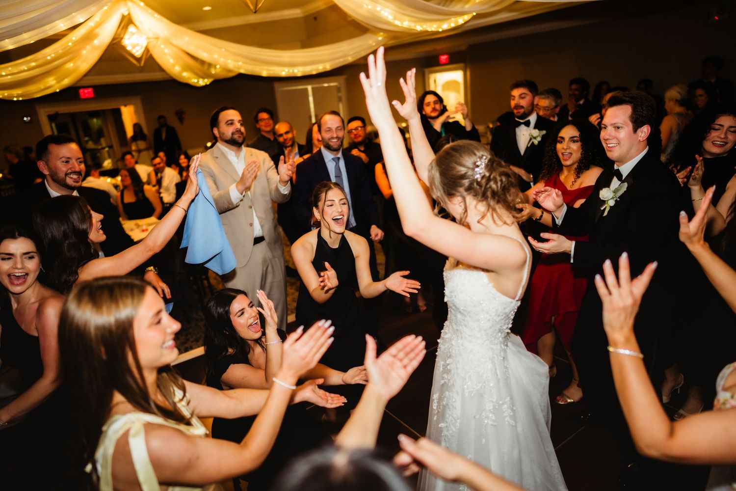 Bride dancing joyfully with guests at a lively wedding reception, surrounded by clapping and cheering attendees.