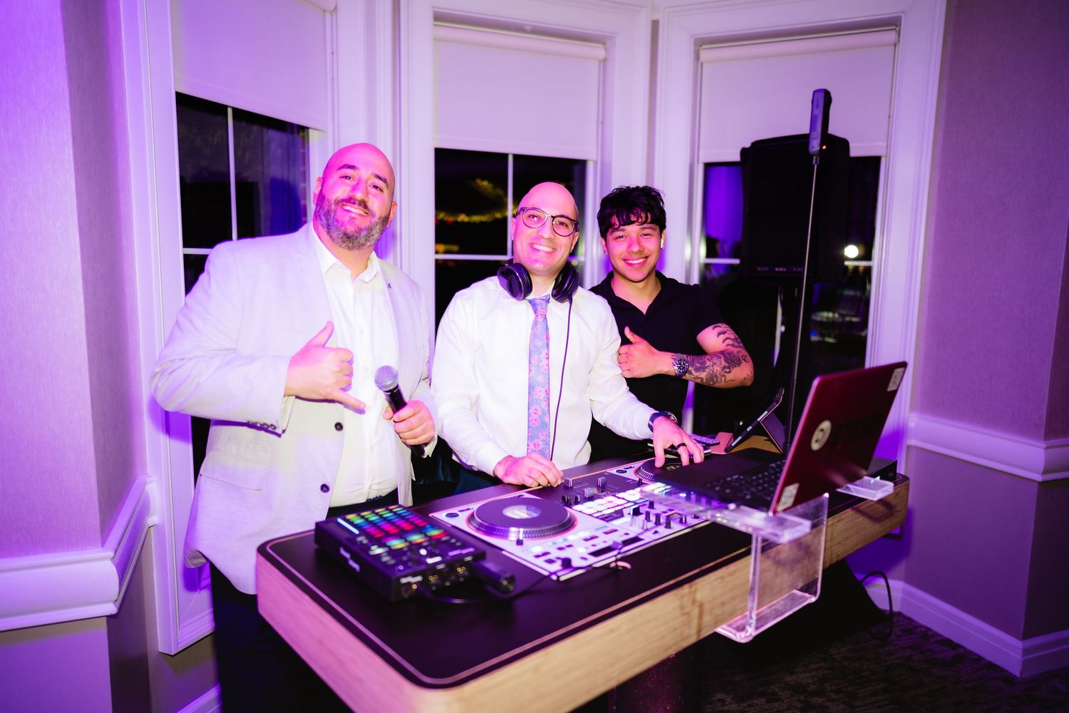 Three men pose behind a DJ booth with equipment at an elegant purple-lit event venue.