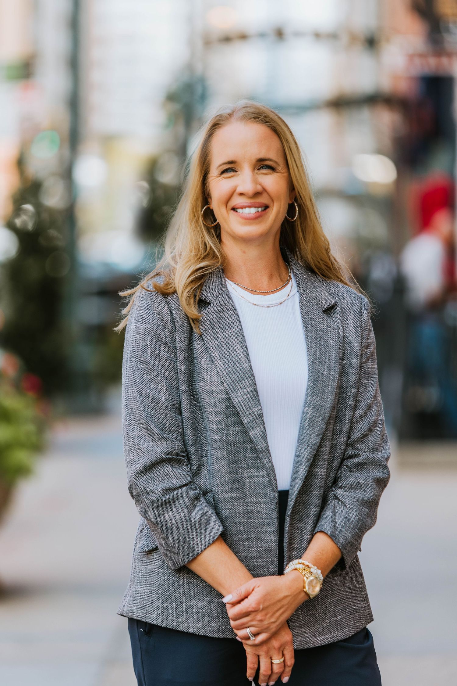 Smiling blonde woman in gray blazer and white top standing on urban street with hands clasped.