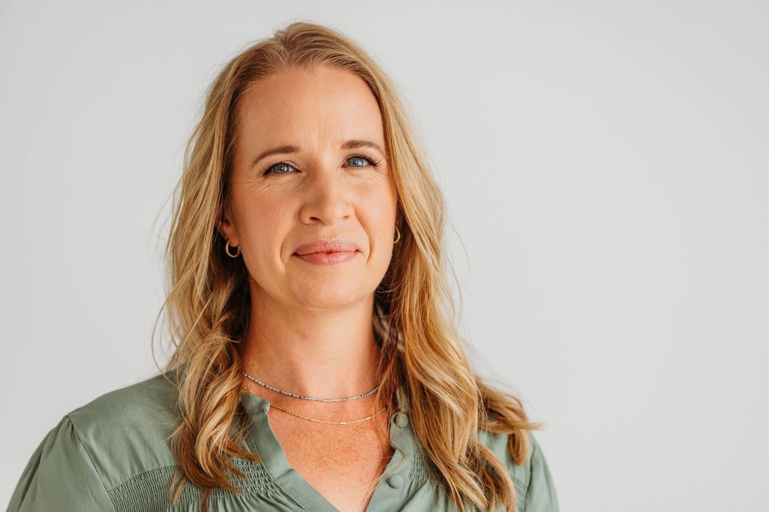Smiling blonde woman in sage green blouse poses for professional headshot against light gray background.