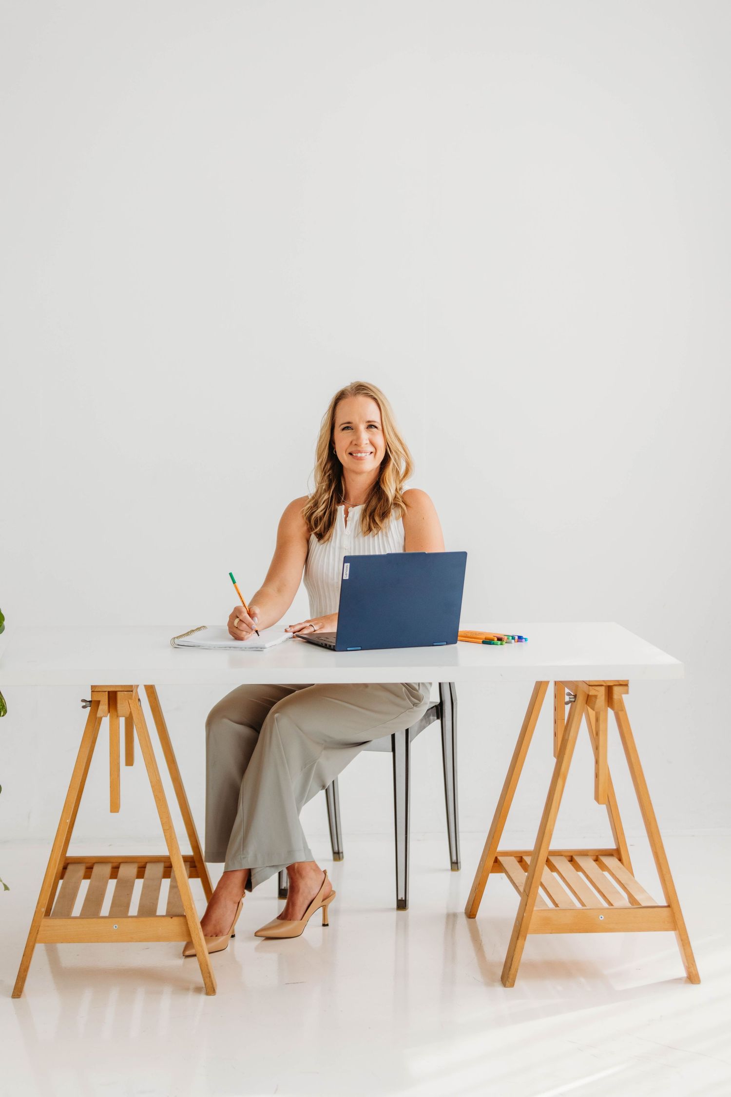 Smiling blonde woman sitting at a white desk with a laptop, writing notes in a bright modern workspace.