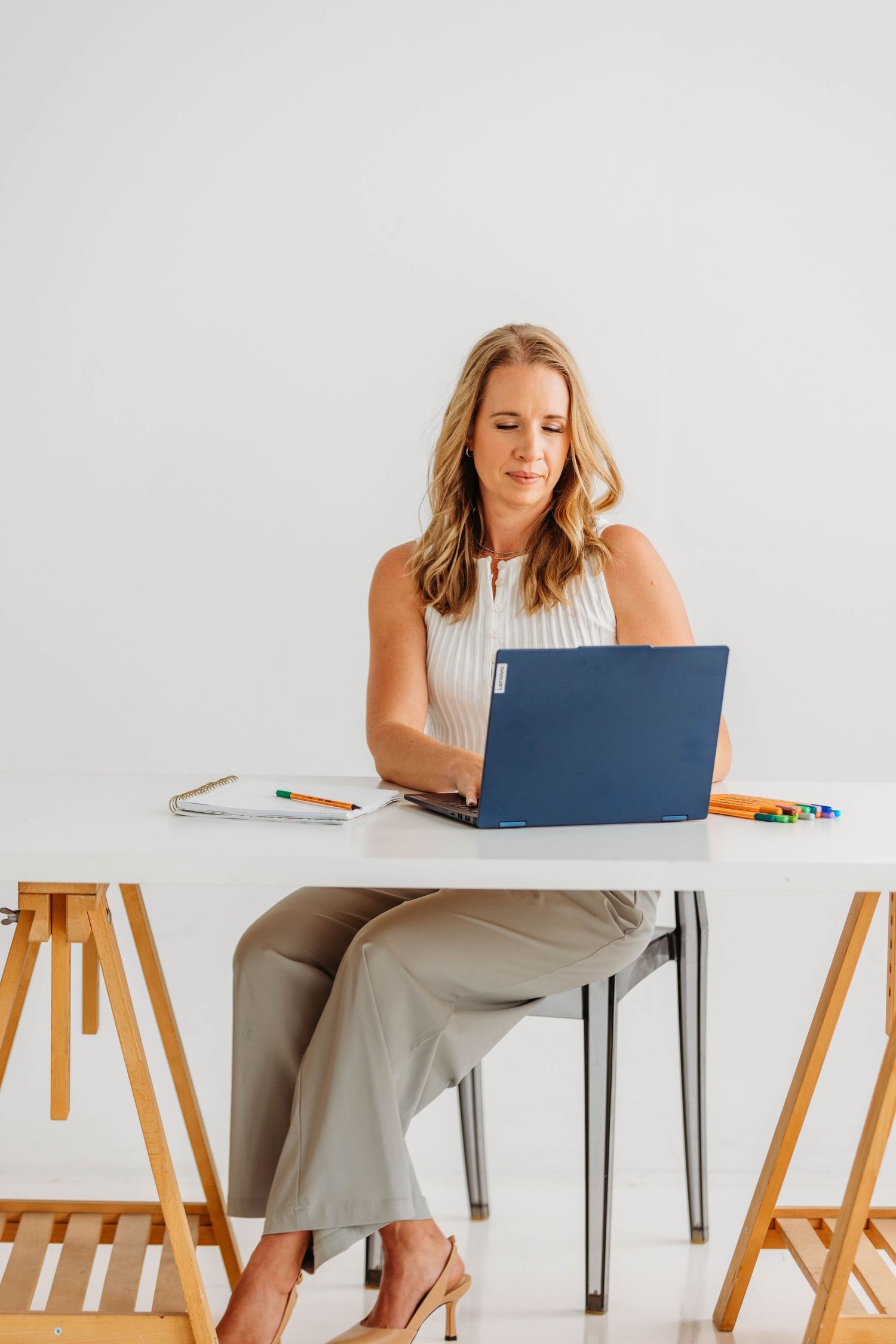 Blonde woman in white sleeveless top and gray pants working on blue laptop at white desk.