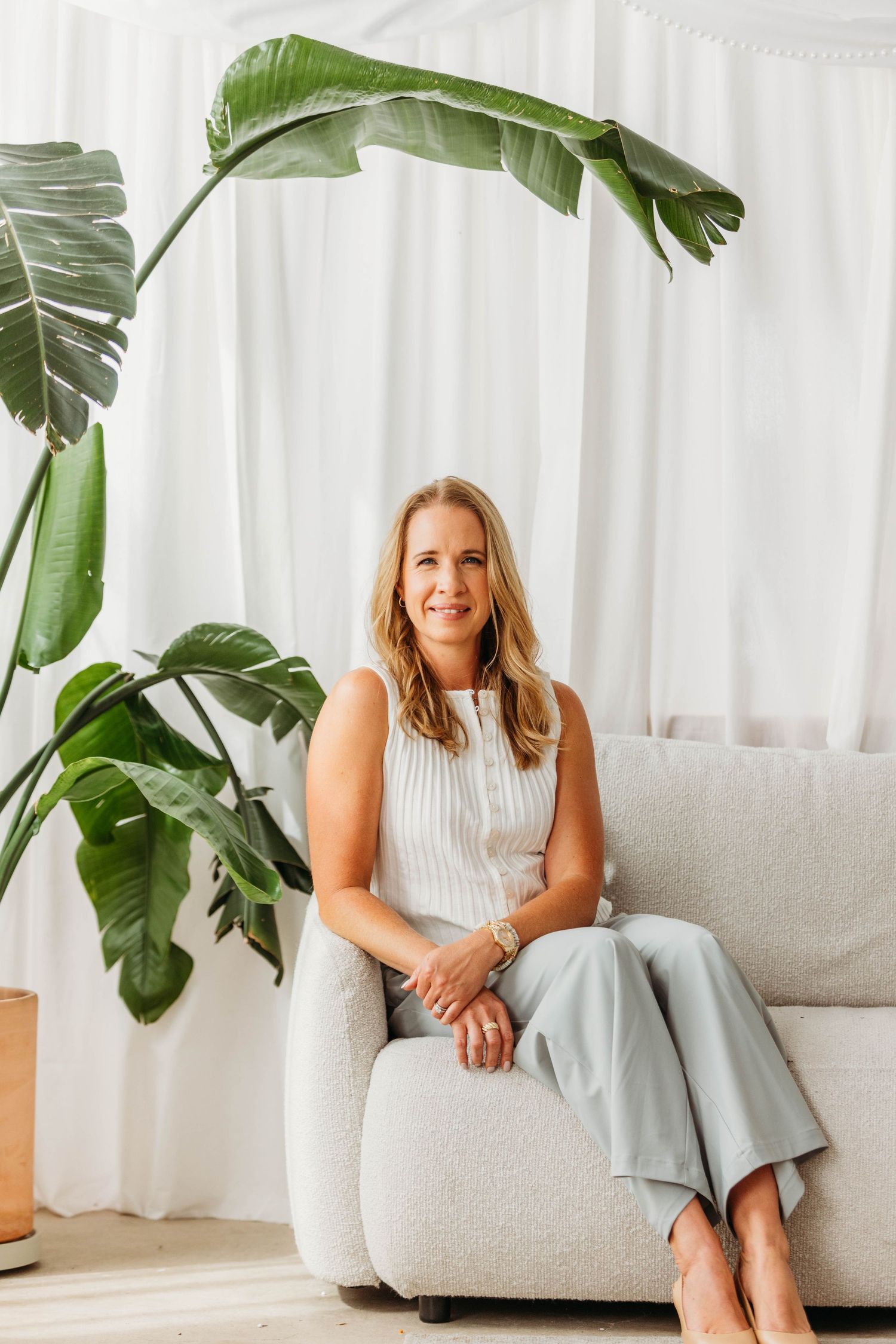 Smiling blonde woman in light blue pants and white top sitting on a cream sofa beside a tropical plant.