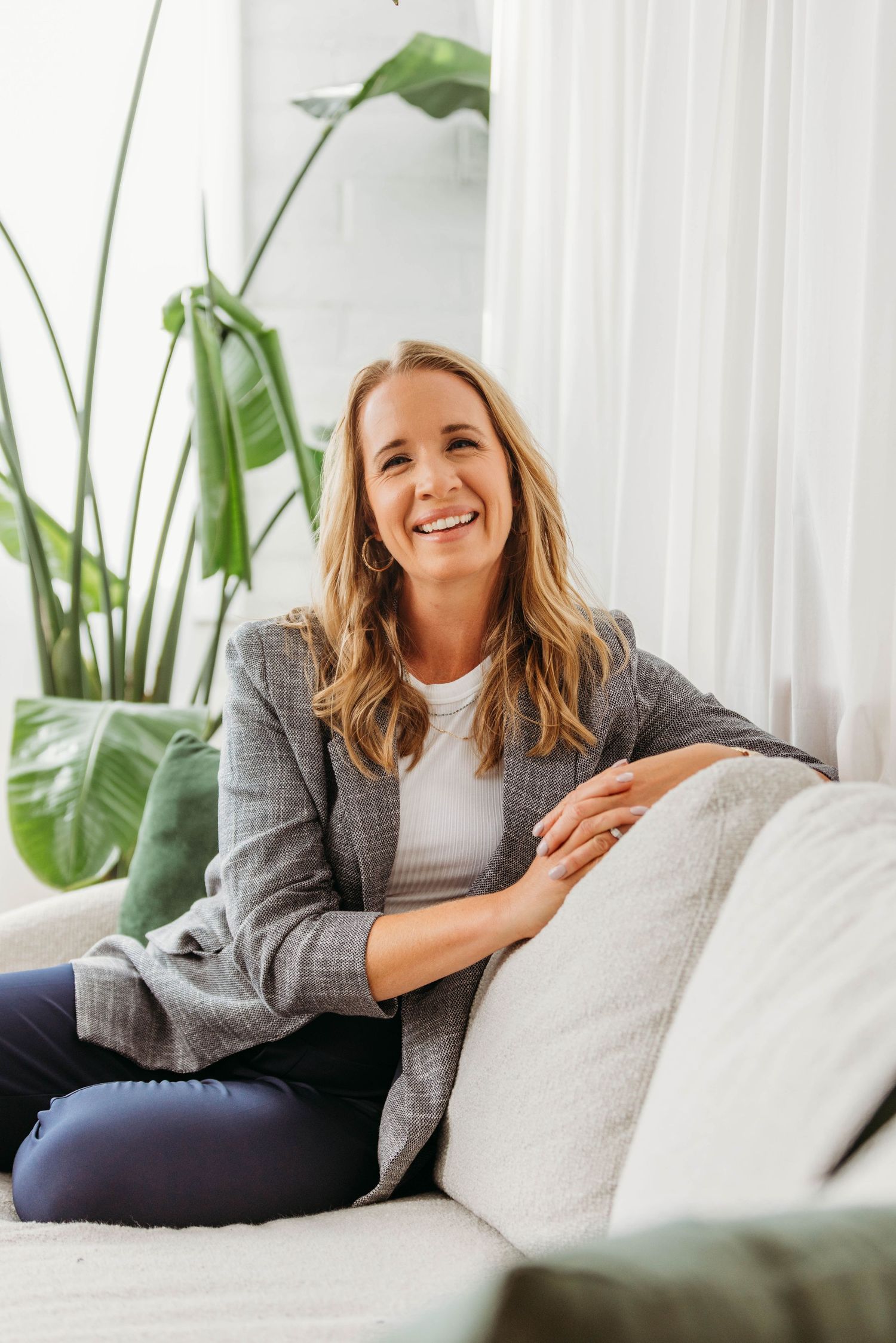 Smiling blonde woman in gray blazer sitting on white sofa with tropical plants in bright living room.