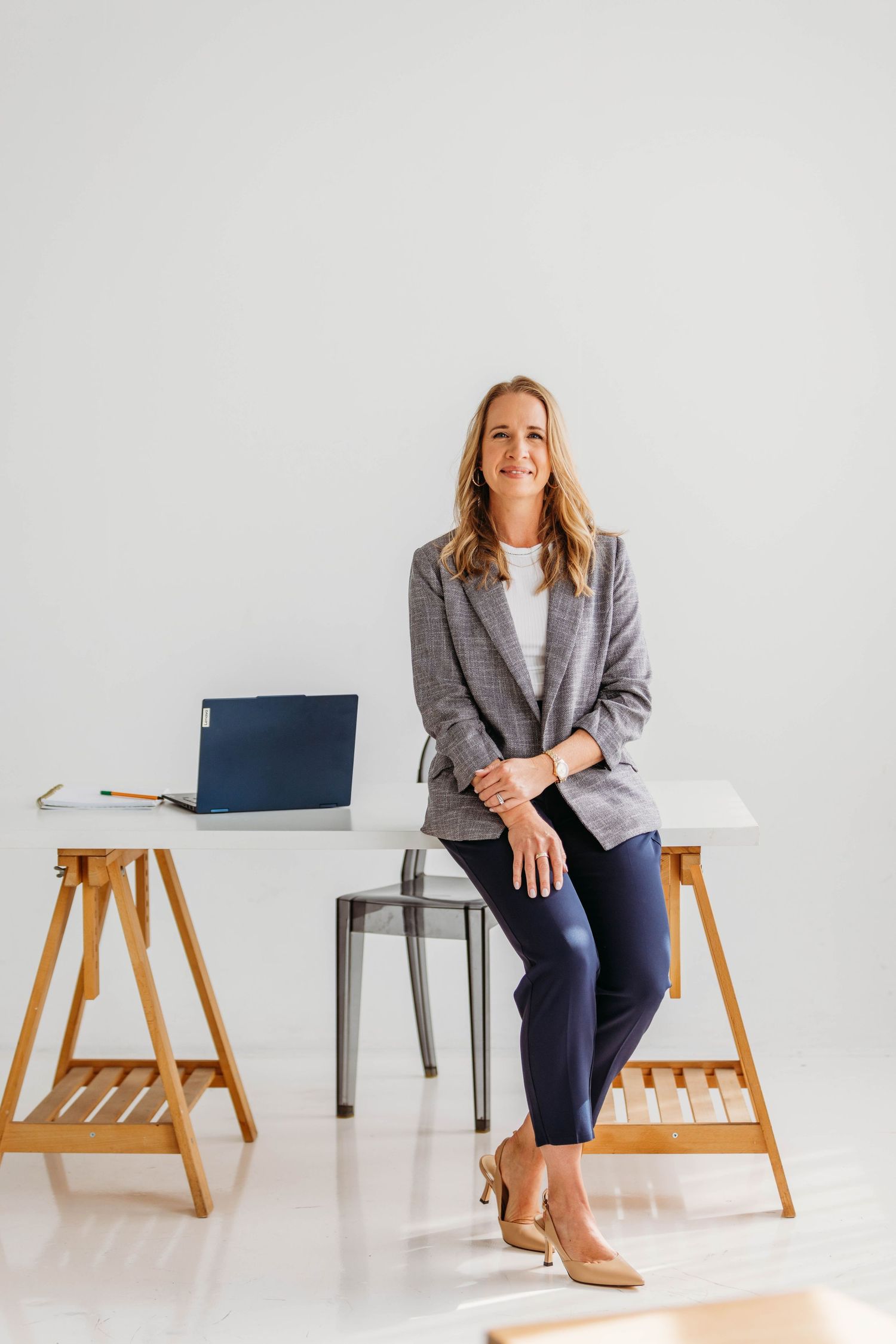 Professional blonde woman in gray blazer leaning against white desk with laptop in bright office.