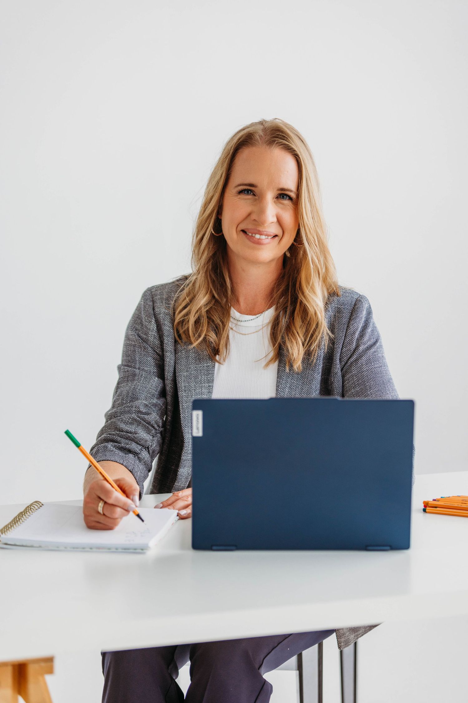 Smiling blonde businesswoman in gray blazer writing notes at white desk with blue laptop in bright office.