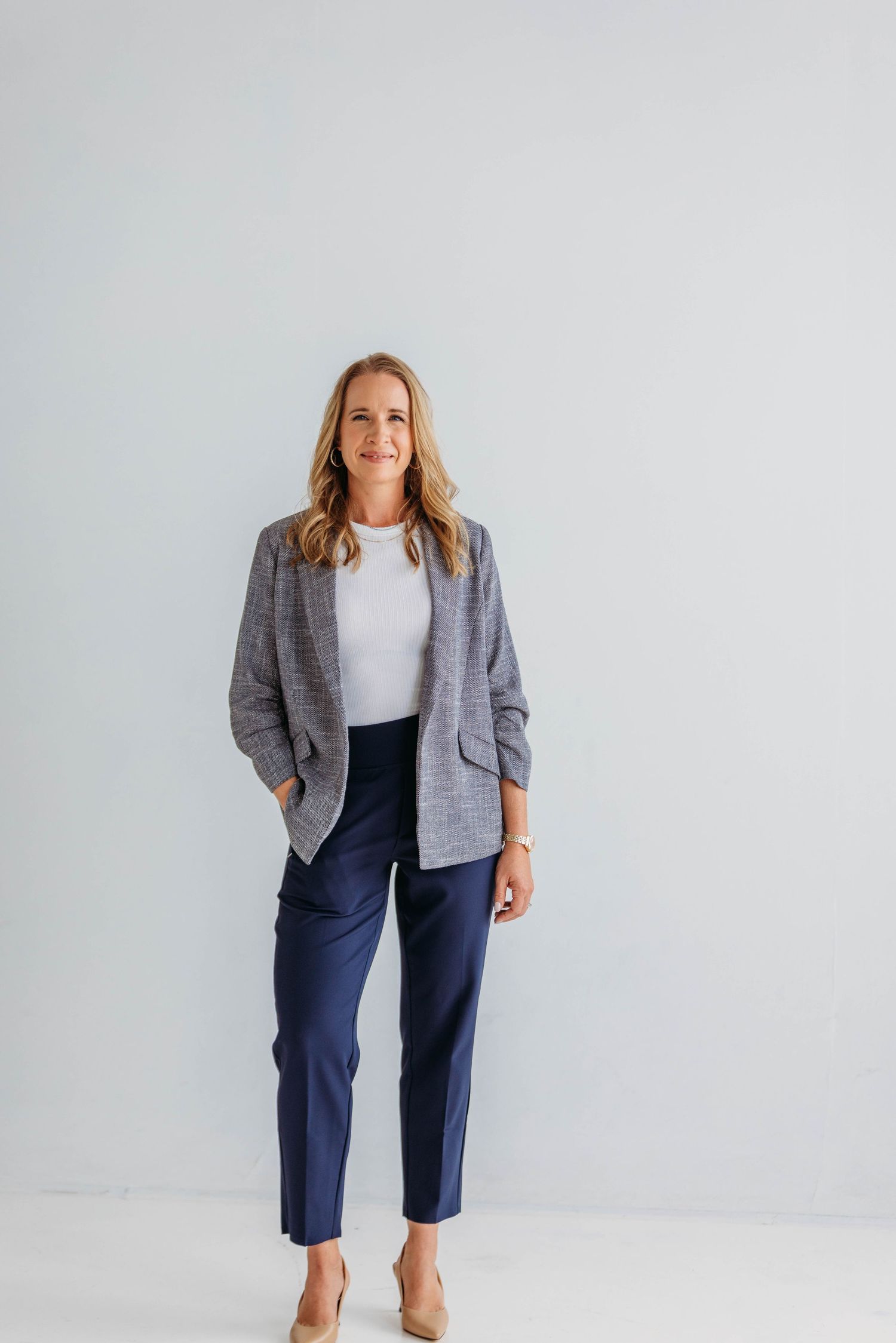 Professional woman in gray blazer, white top, and navy pants standing confidently against a light gray background.
