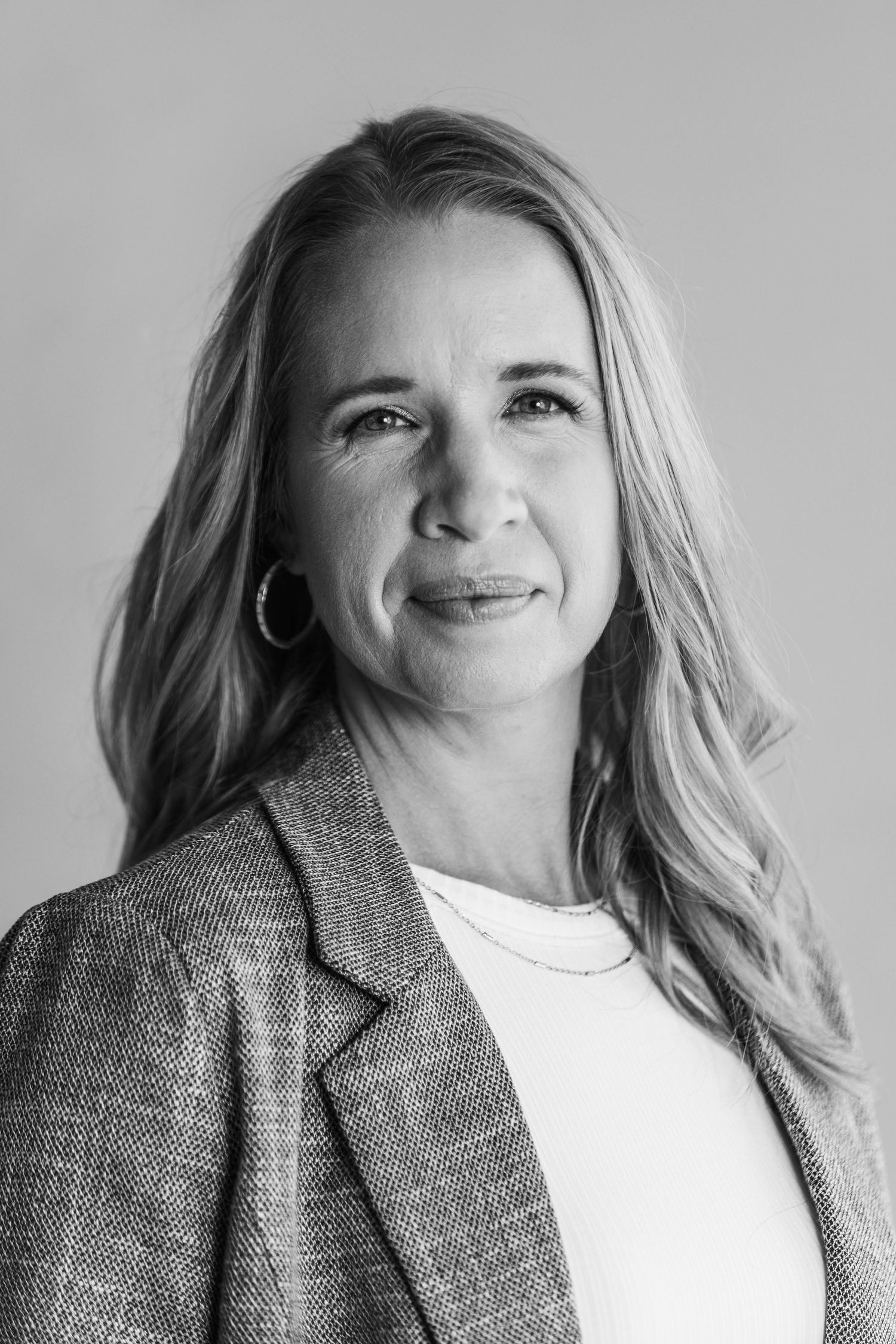 Black and white professional headshot of a blonde woman in a blazer and hoop earrings against a neutral background.