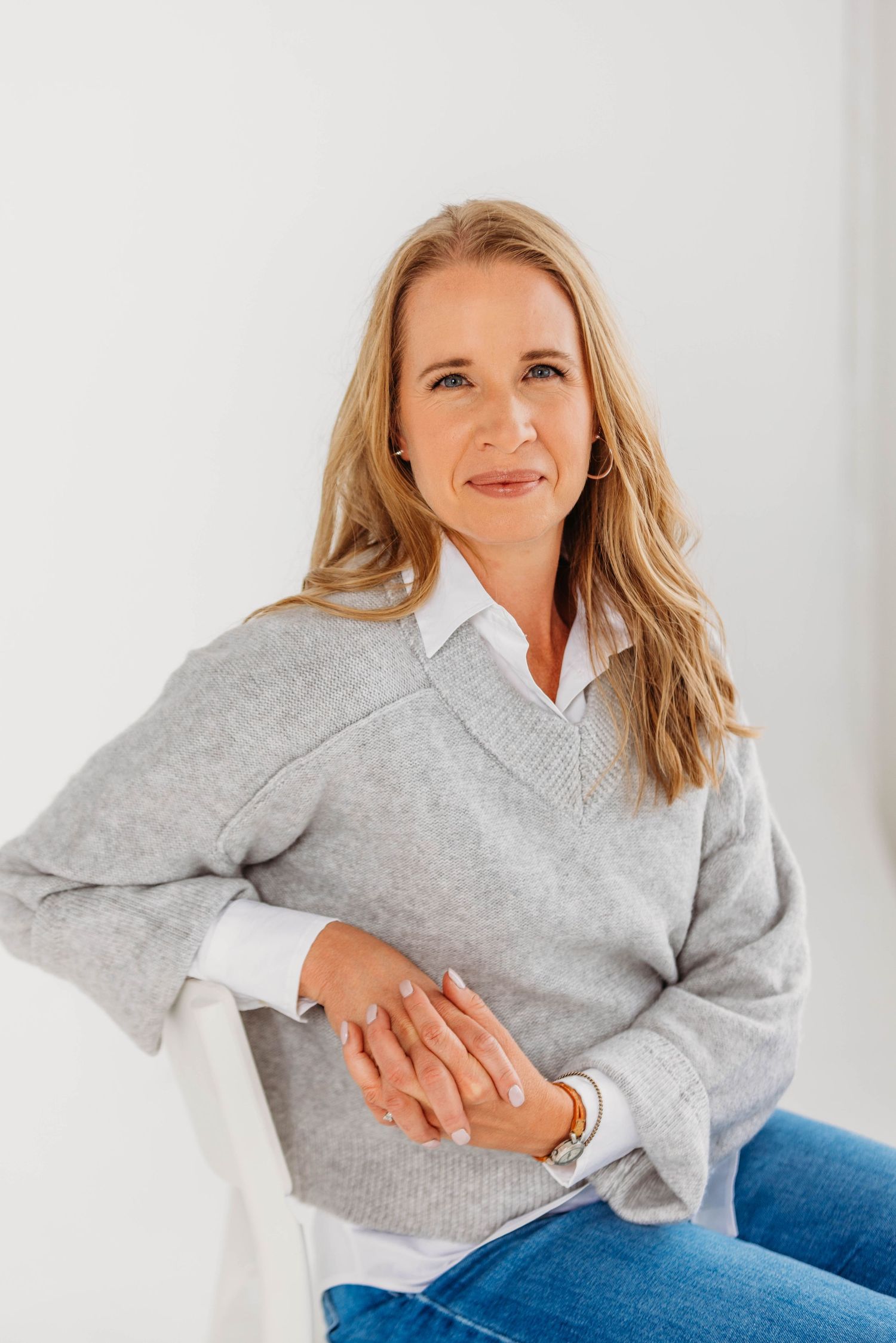 Confident blonde woman in gray sweater and jeans sitting relaxed on white chair against light background.