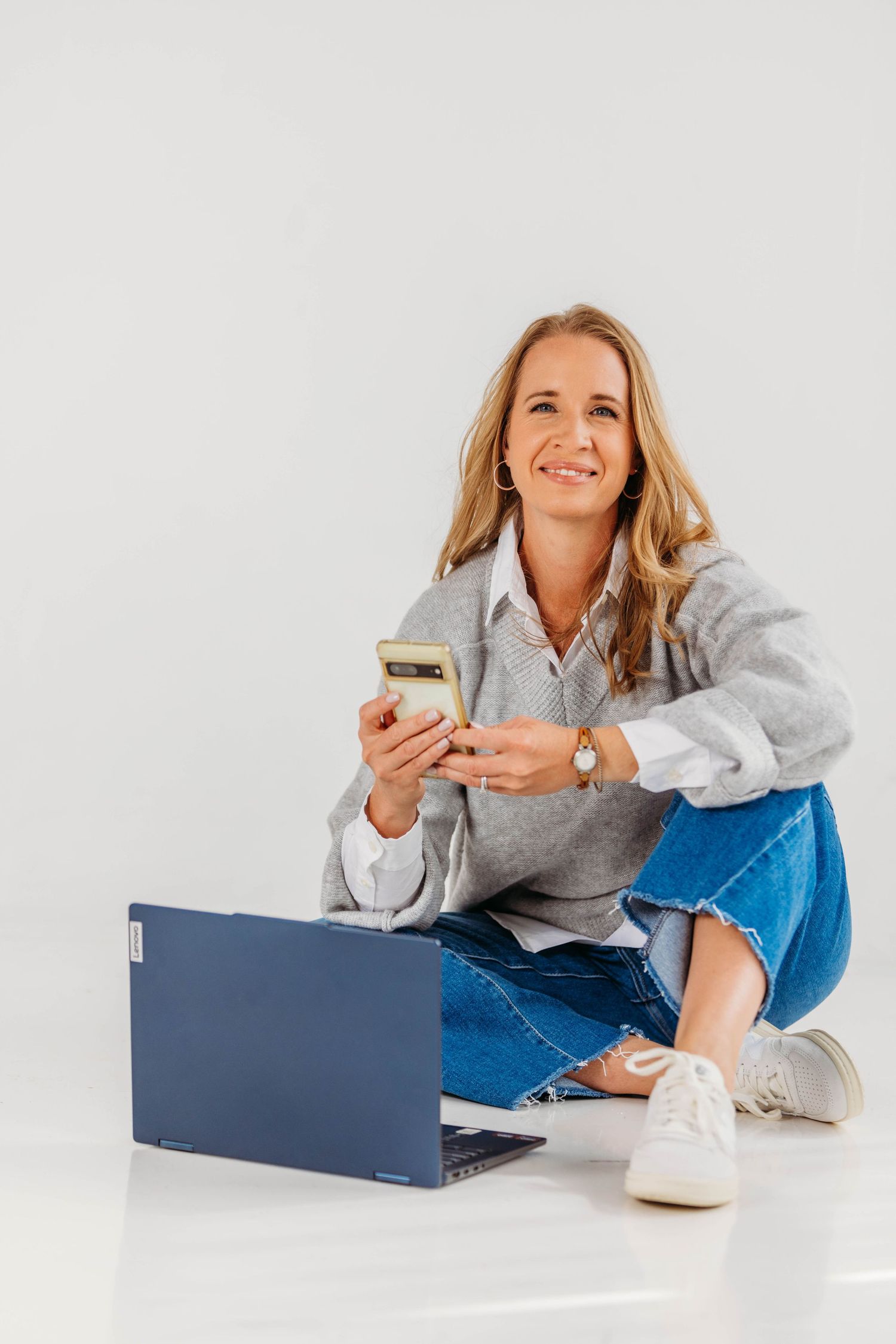 Smiling blonde woman sitting on floor with laptop and smartphone in casual gray sweater and jeans.