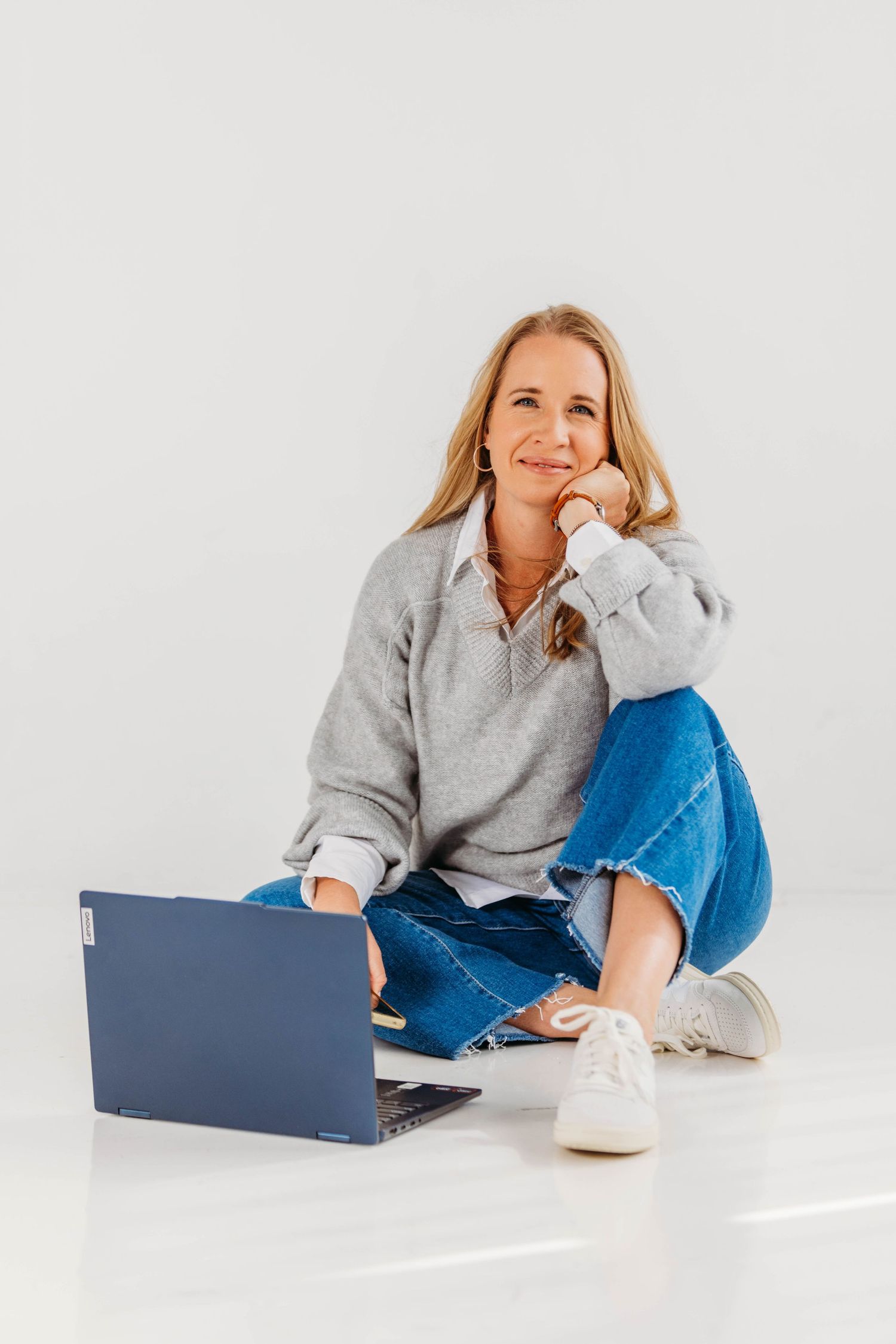 Casual blonde woman in gray hoodie and jeans sitting cross-legged on floor with blue laptop nearby.