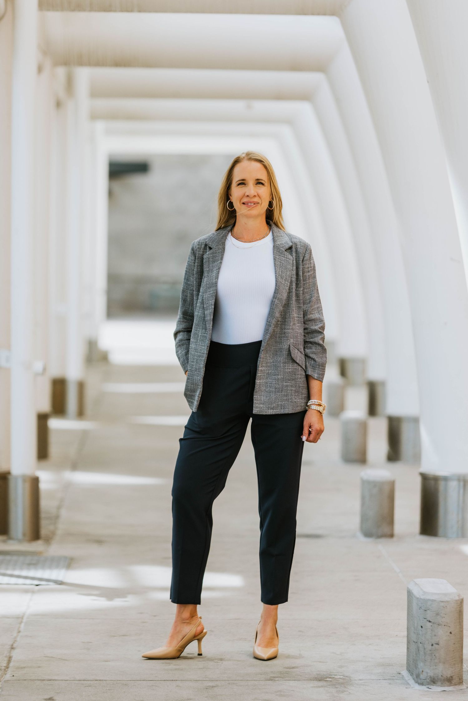 Professional woman in gray blazer, white top, and black trousers posing in modern white colonnade architecture.