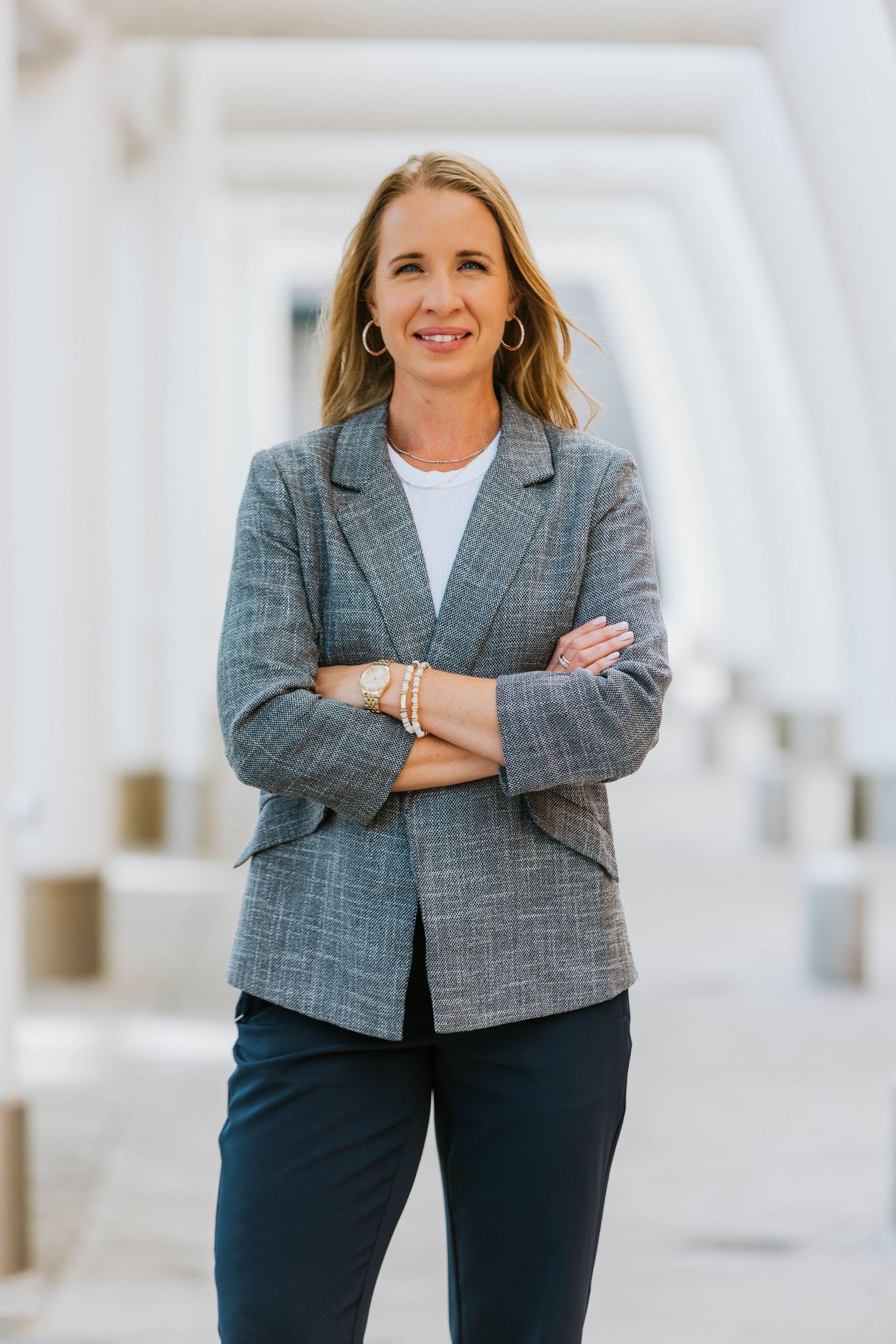 Professional woman in gray blazer stands confidently with arms crossed in a bright white columned hallway.