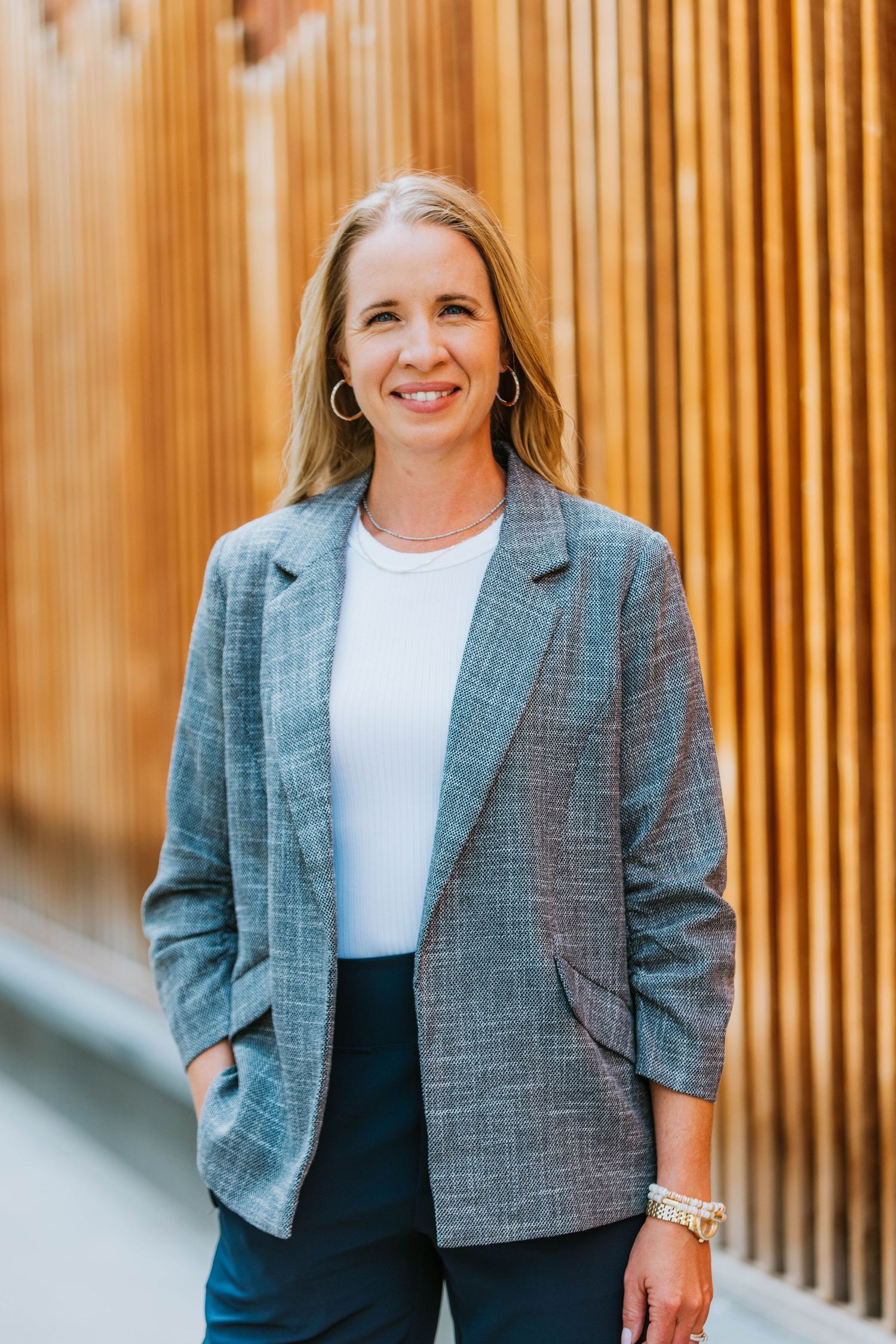 Professional woman in gray blazer and white top smiling confidently in modern office corridor.