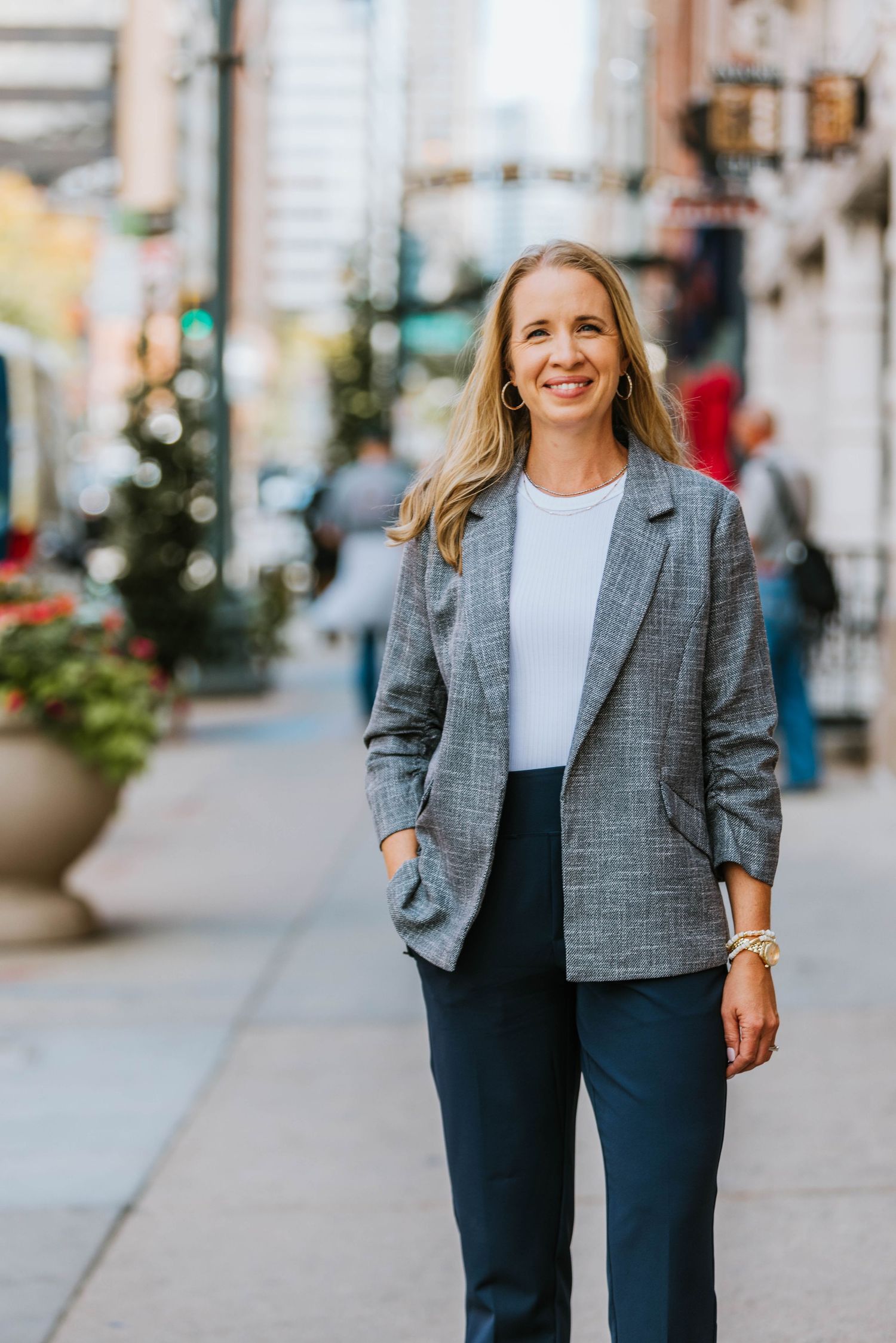 Smiling businesswoman in gray blazer and navy pants standing on a city sidewalk in an urban setting.