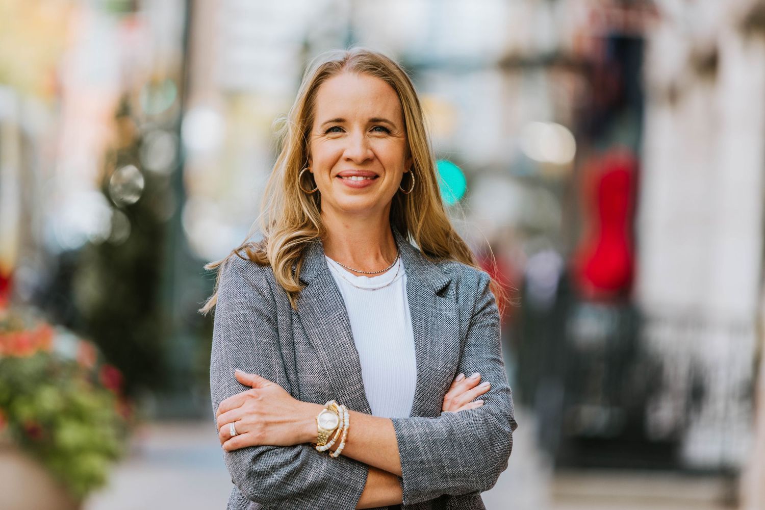 Professional woman in gray blazer smiling with arms crossed on a vibrant city street.