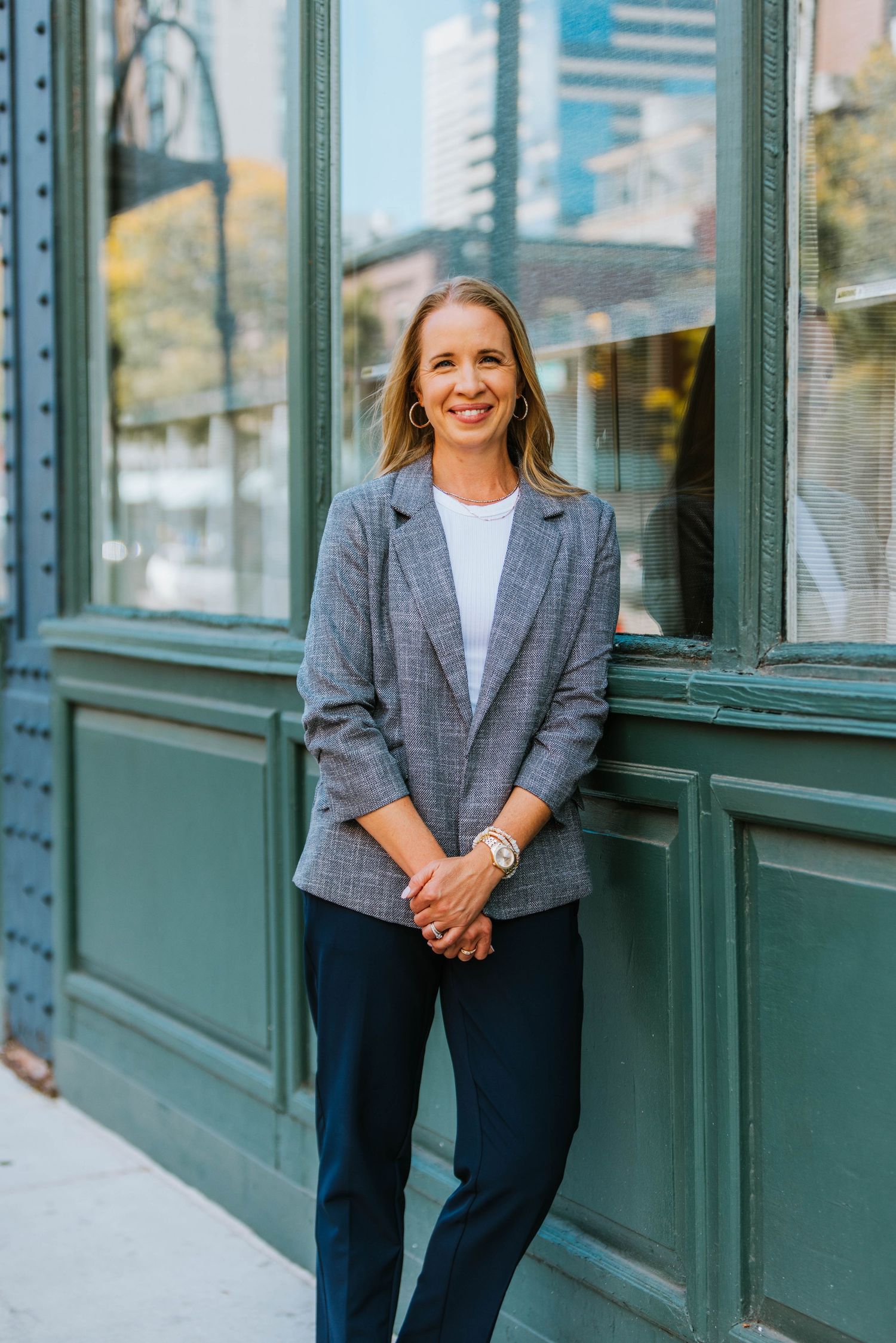 Professional woman in gray blazer smiling outdoors leaning against green building facade in urban setting.