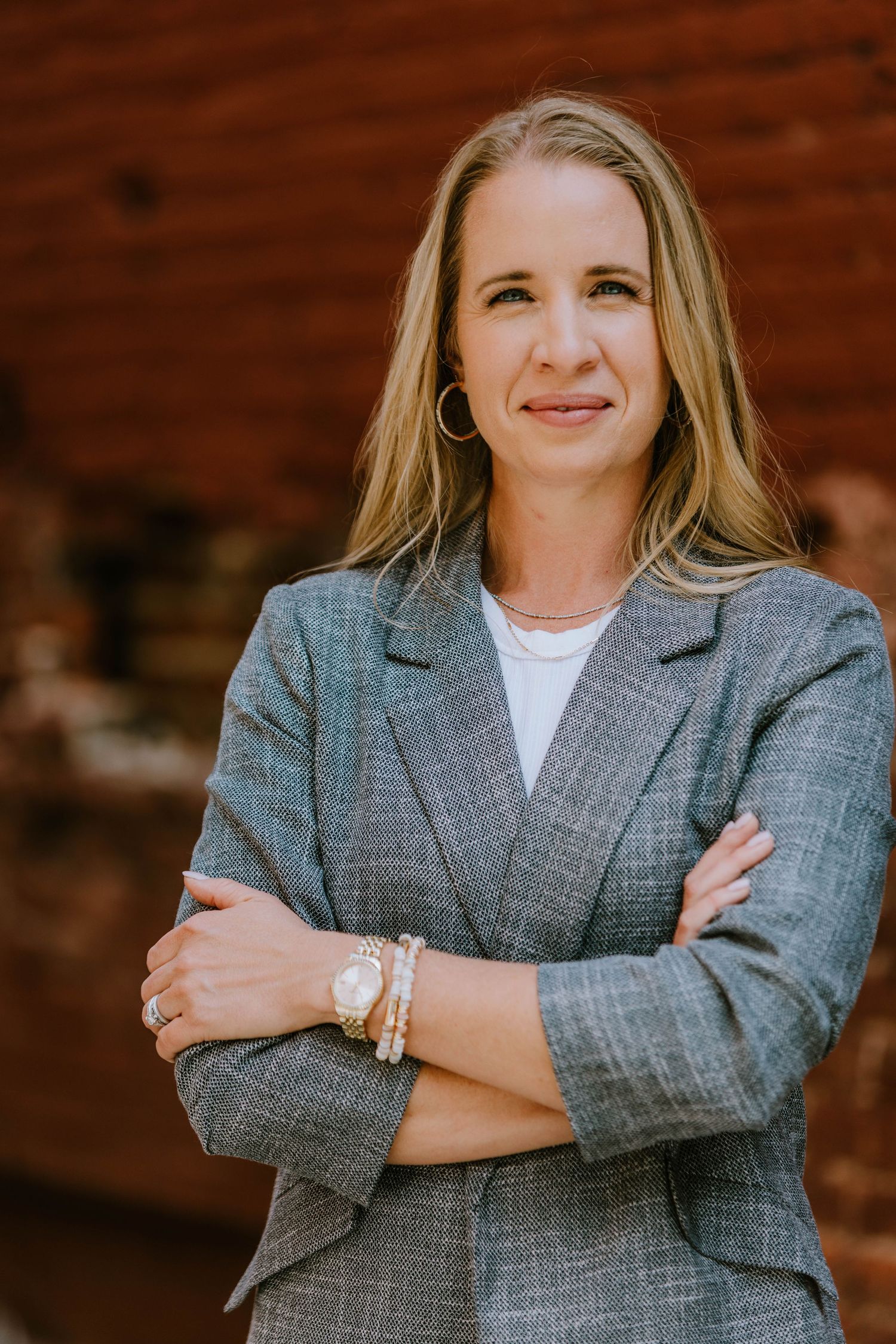 Professional woman in gray blazer with arms crossed, smiling confidently outdoors against a brick wall background.