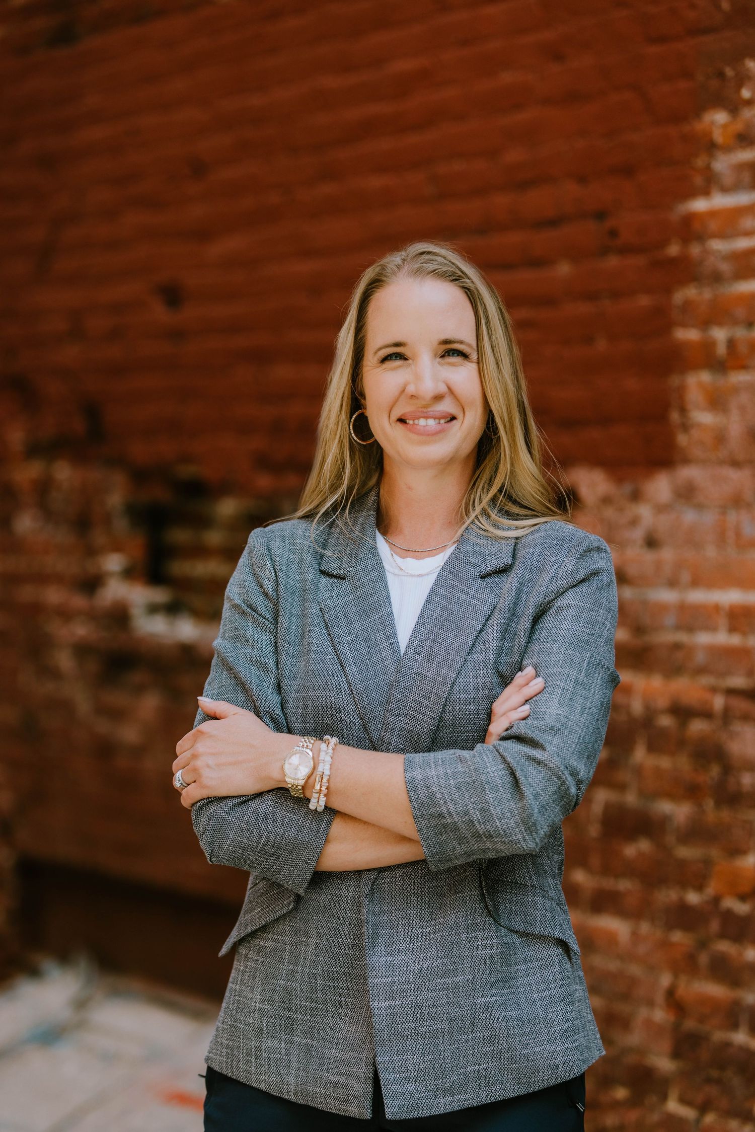 Confident blonde woman in gray blazer posing with arms crossed against rustic brick wall backdrop.