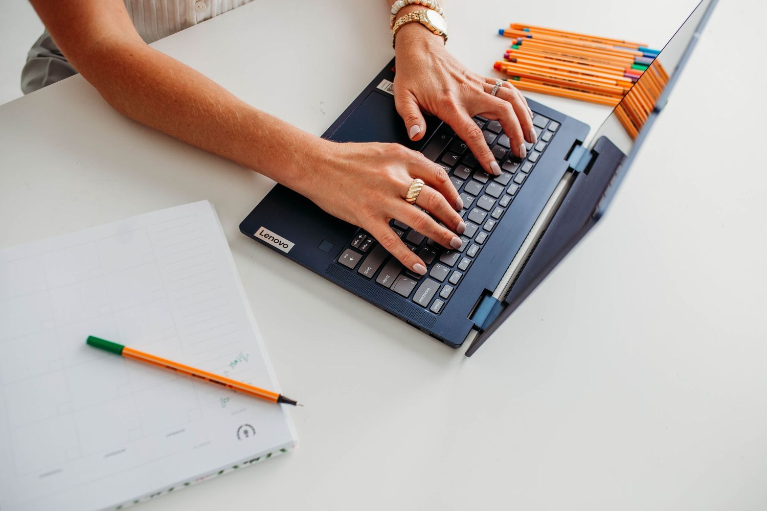 Woman typing on a laptop at a white desk with pencils and a notebook, working in a bright workspace.