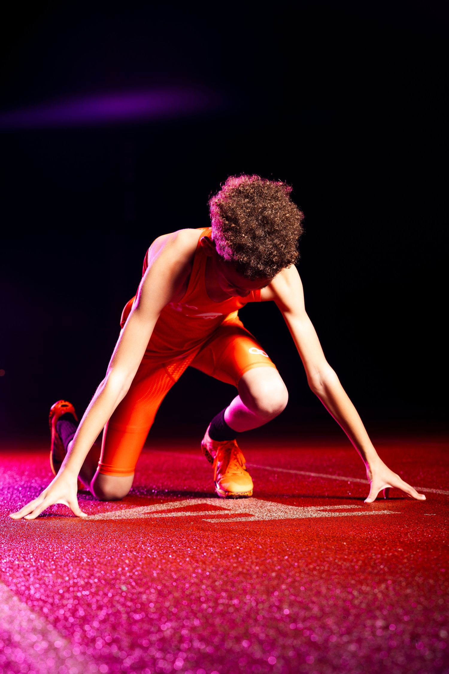 A sprinter in an orange uniform crouches in starting position on a red track under dramatic pink and purple lighting.