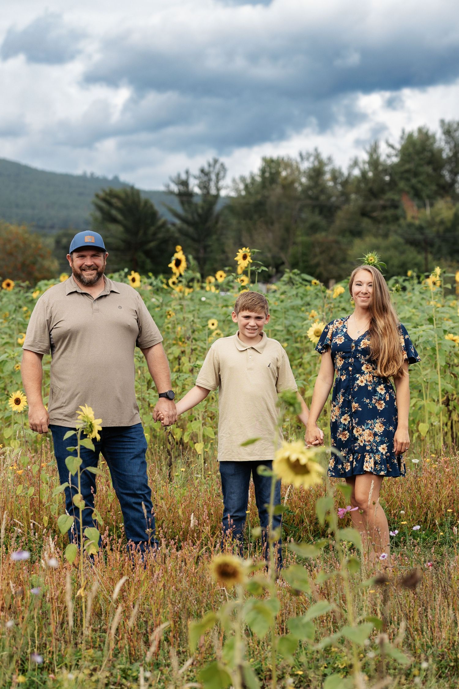 Family of three holding hands in a sunflower field with mountains and trees in the background on a cloudy day.