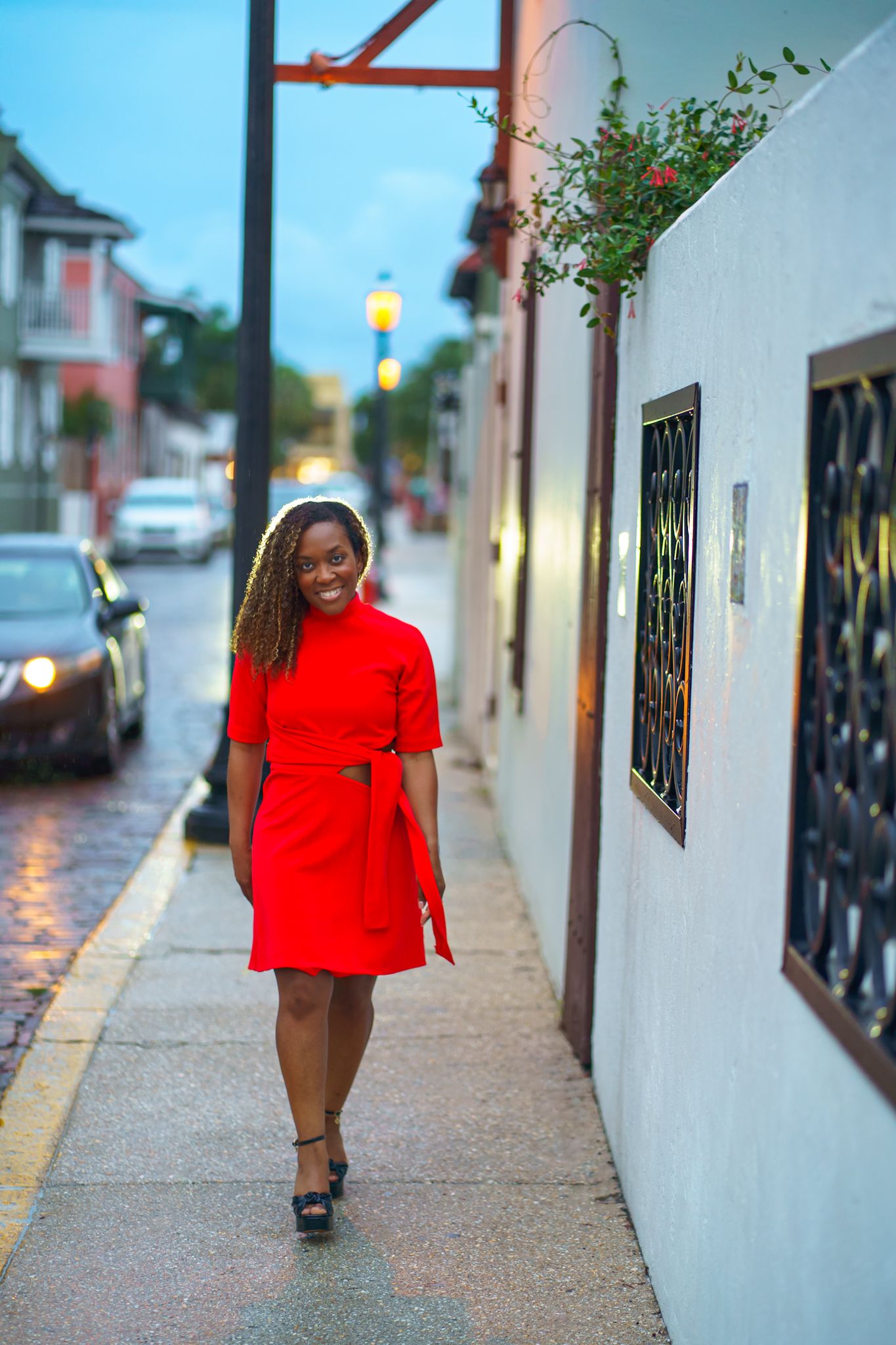 A smiling woman in a red cut-out dress walks along a wet urban sidewalk at dusk near a white building.