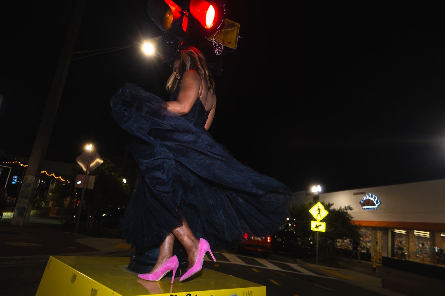 Woman in blue gown and pink heels standing on yellow barrier under red traffic light at night.