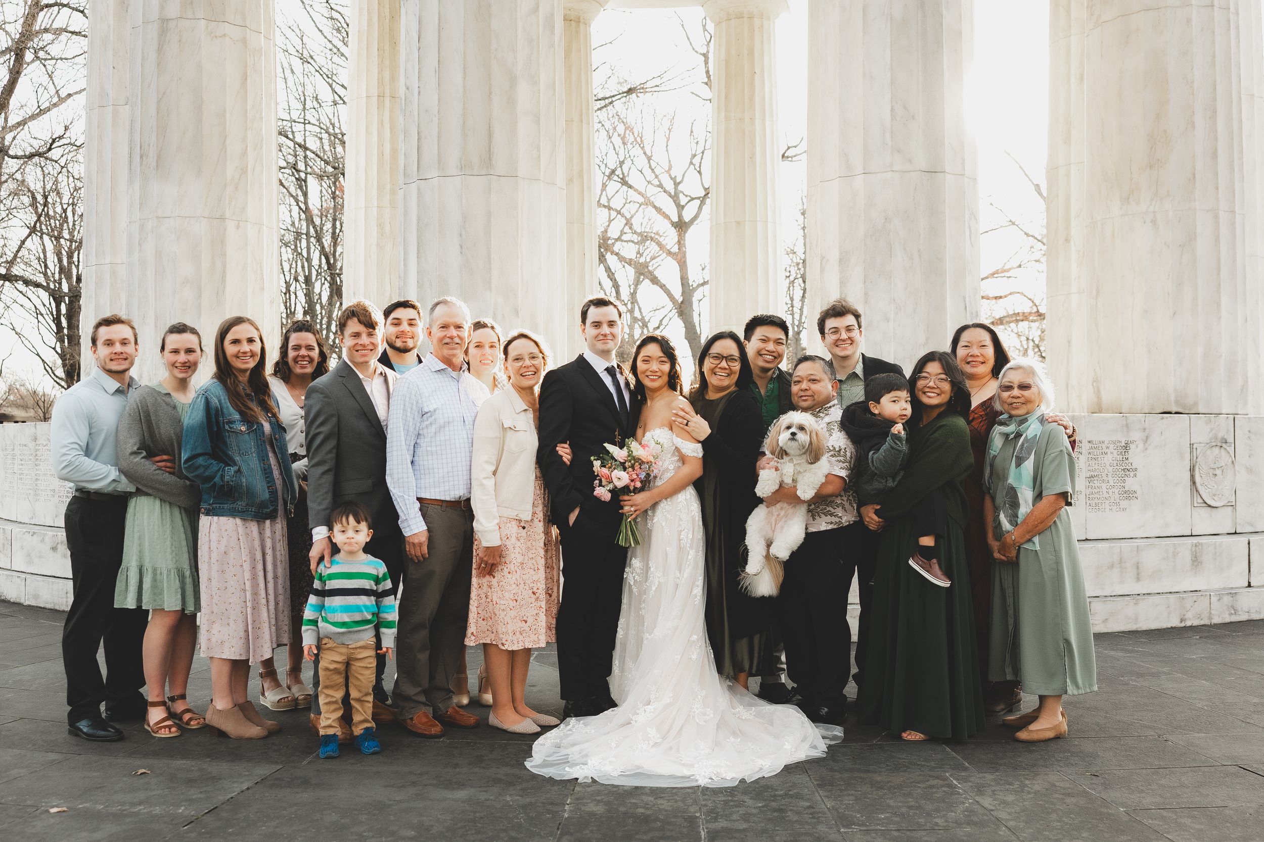 Wedding party poses with bride and groom before grand white columns, including family members and a fluffy dog.
