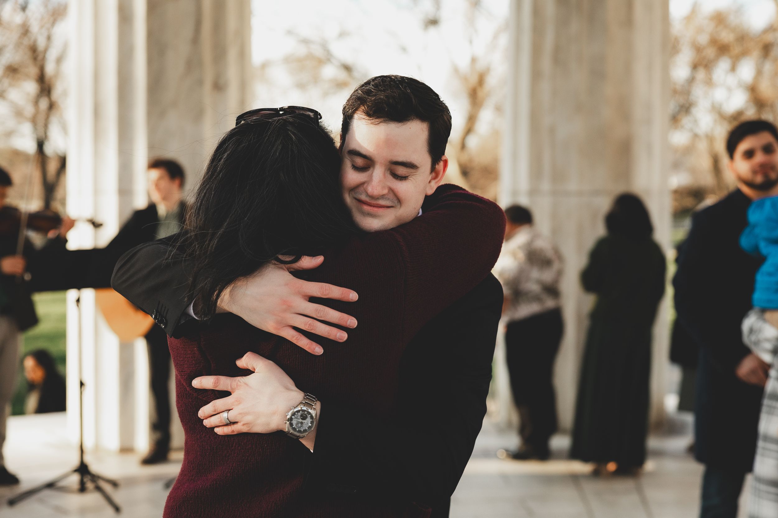 A smiling groom hugs a guest at an outdoor wedding ceremony near elegant stone columns.