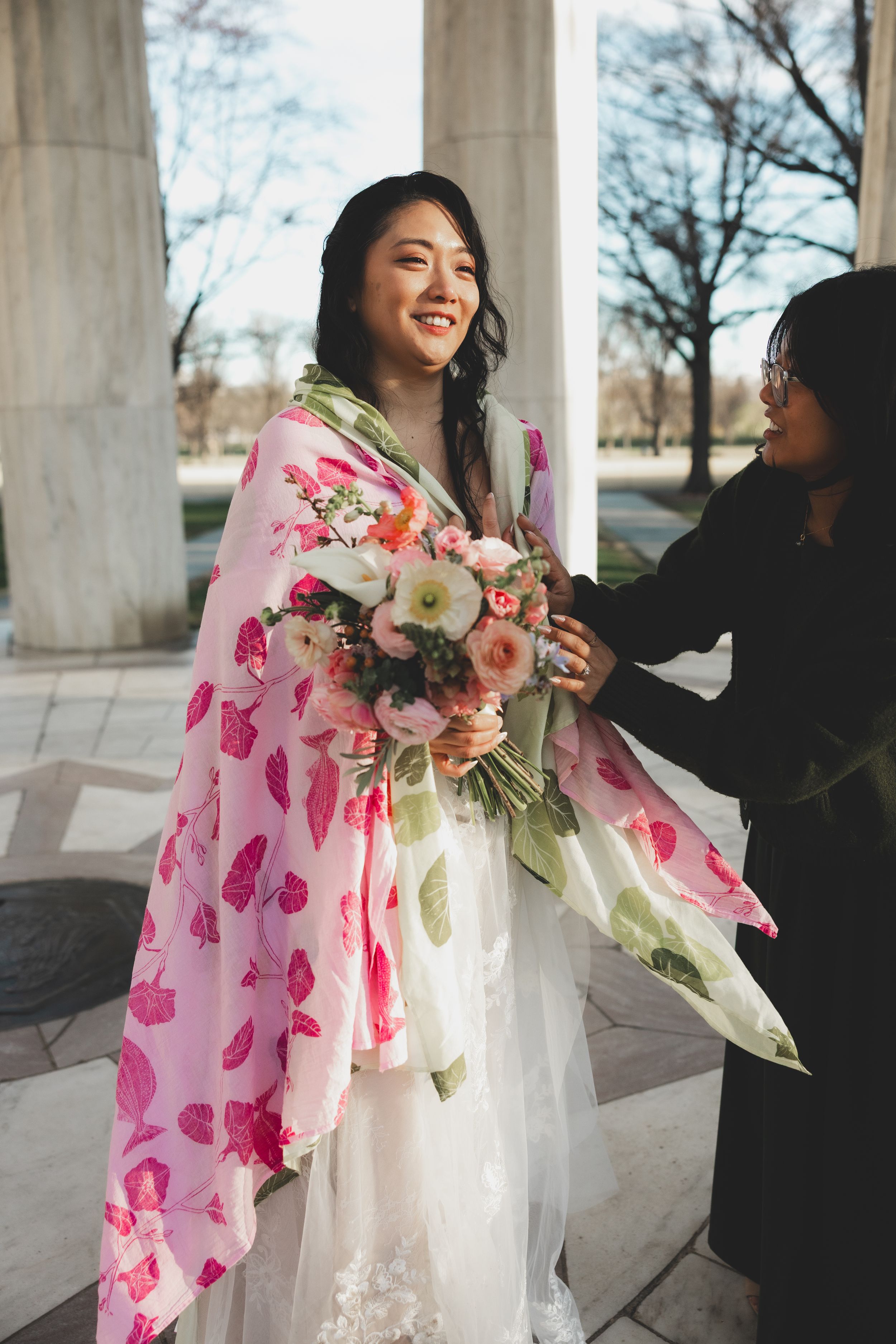 A smiling bride in a white gown with a pink floral wrap holds a bouquet at an outdoor wedding.
