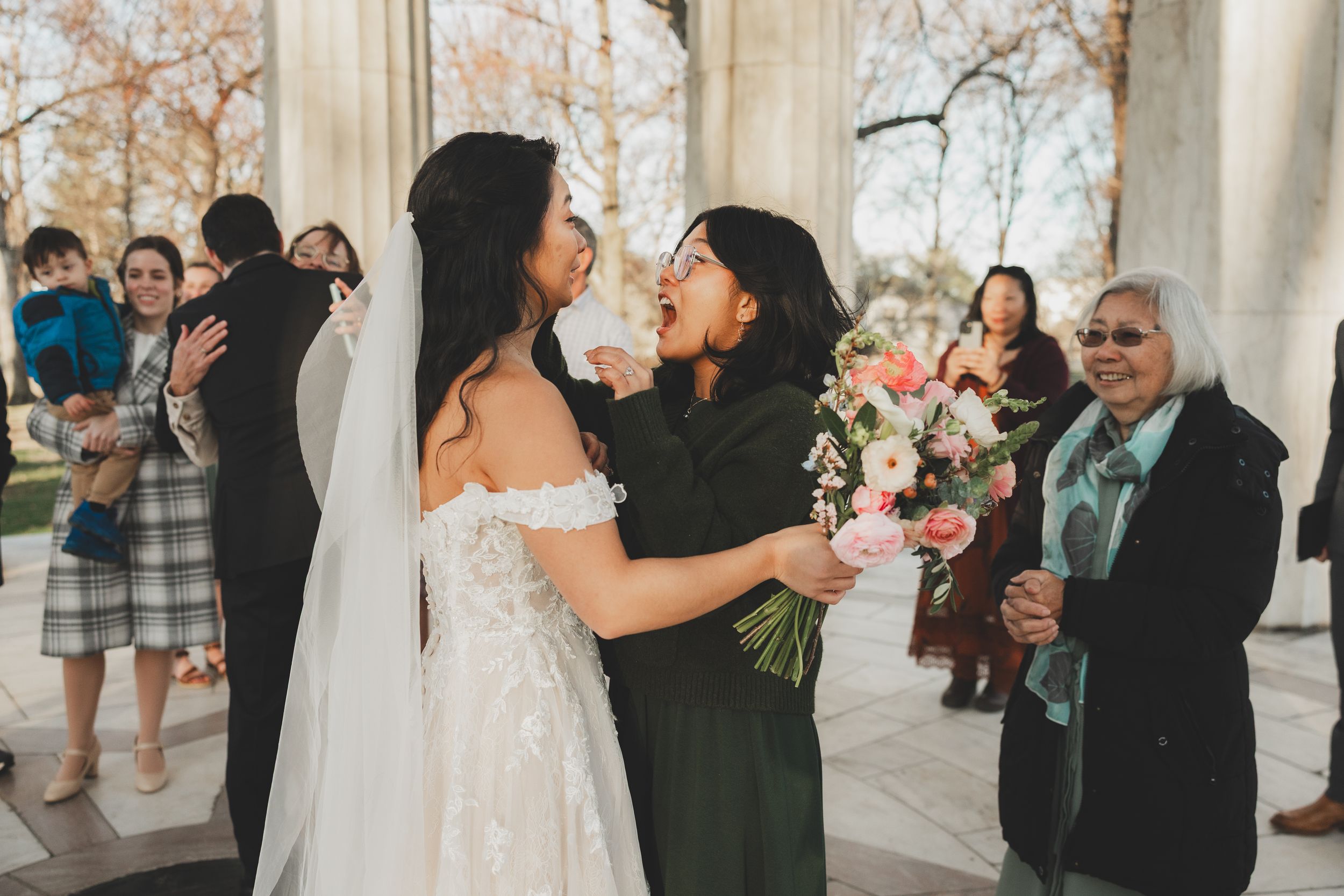 Bride in lace wedding gown receives warm congratulations from guests at an outdoor wedding ceremony.