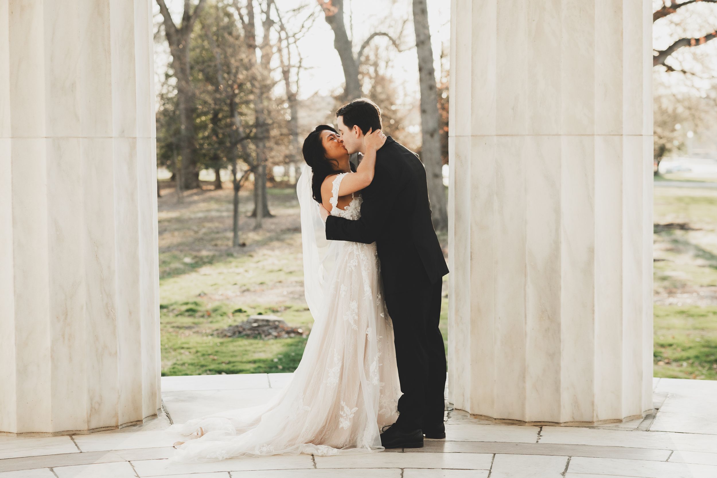A bride and groom share a romantic kiss between white columns inside the DC War Memorial in Washington D.C.