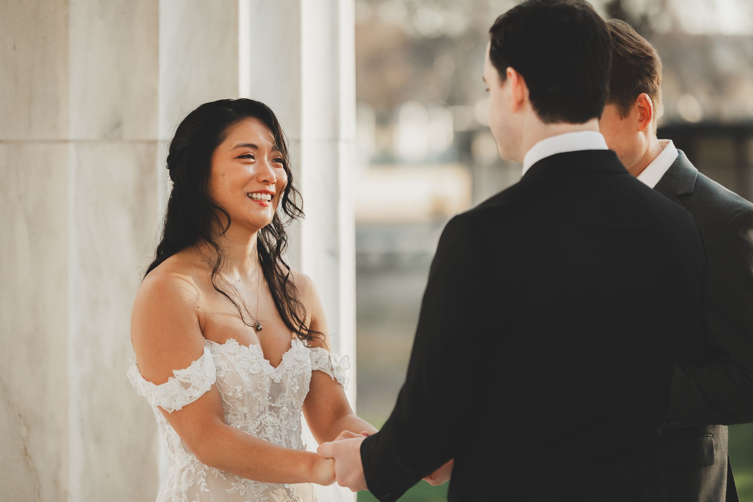 Bride in off-shoulder lace gown smiling joyfully at groom in black tuxedo during outdoor wedding ceremony inside the dc WWII memorial