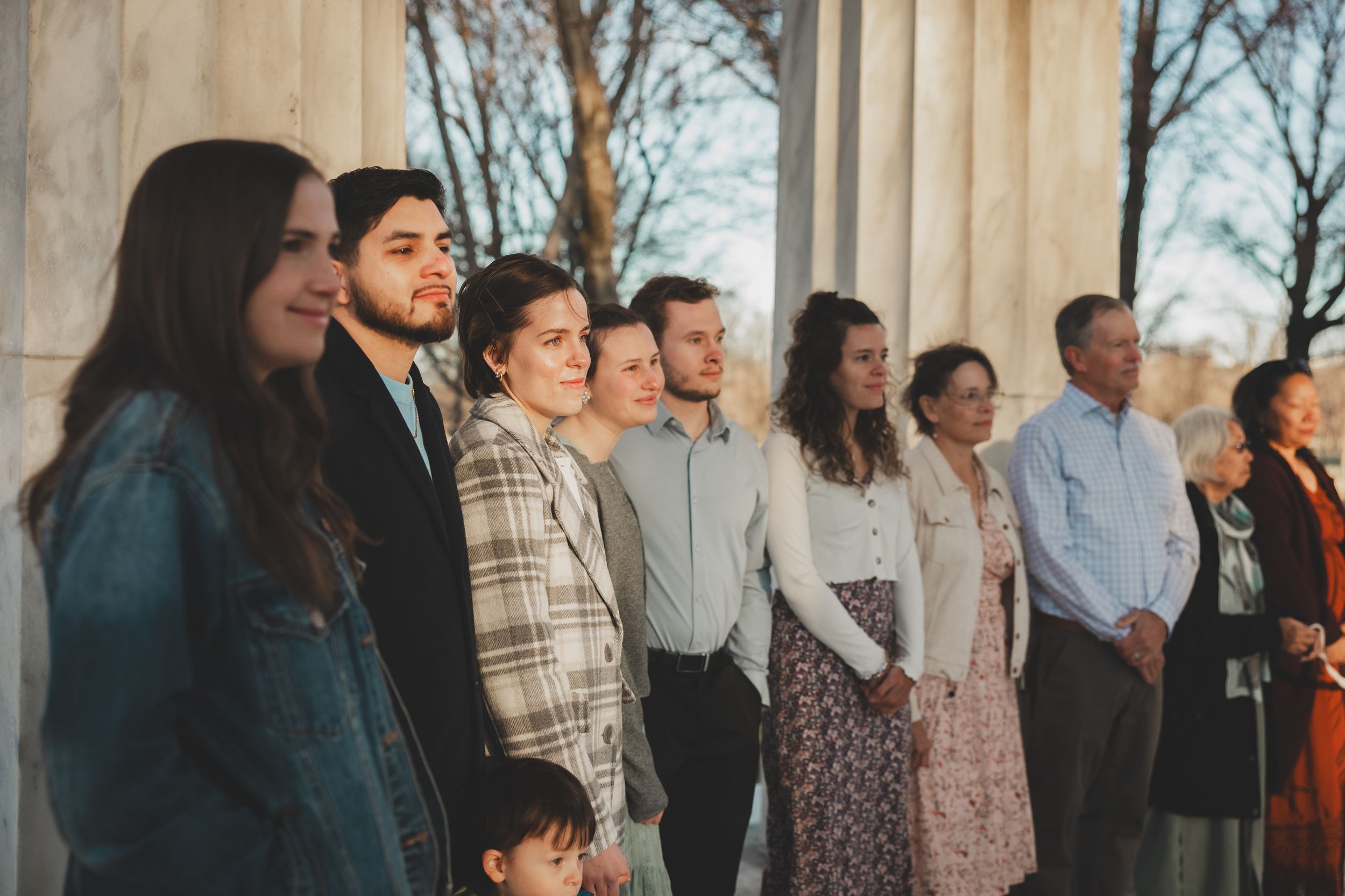 Group of elegantly dressed wedding guests standing together between marble columns at an outdoor ceremony.